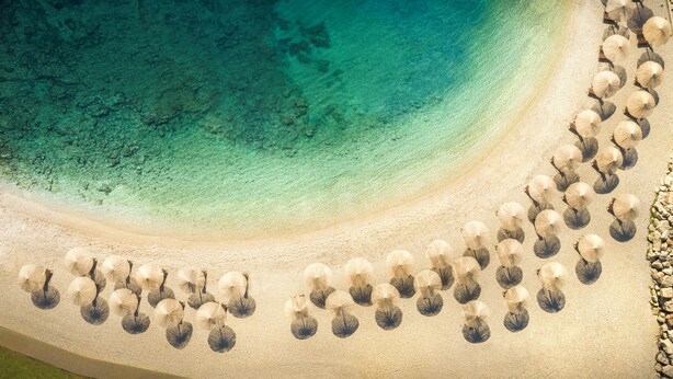 Vista aérea de uma praia com guarda-sóis dispostos em círculo e um corpo d'água azul cristalino.
