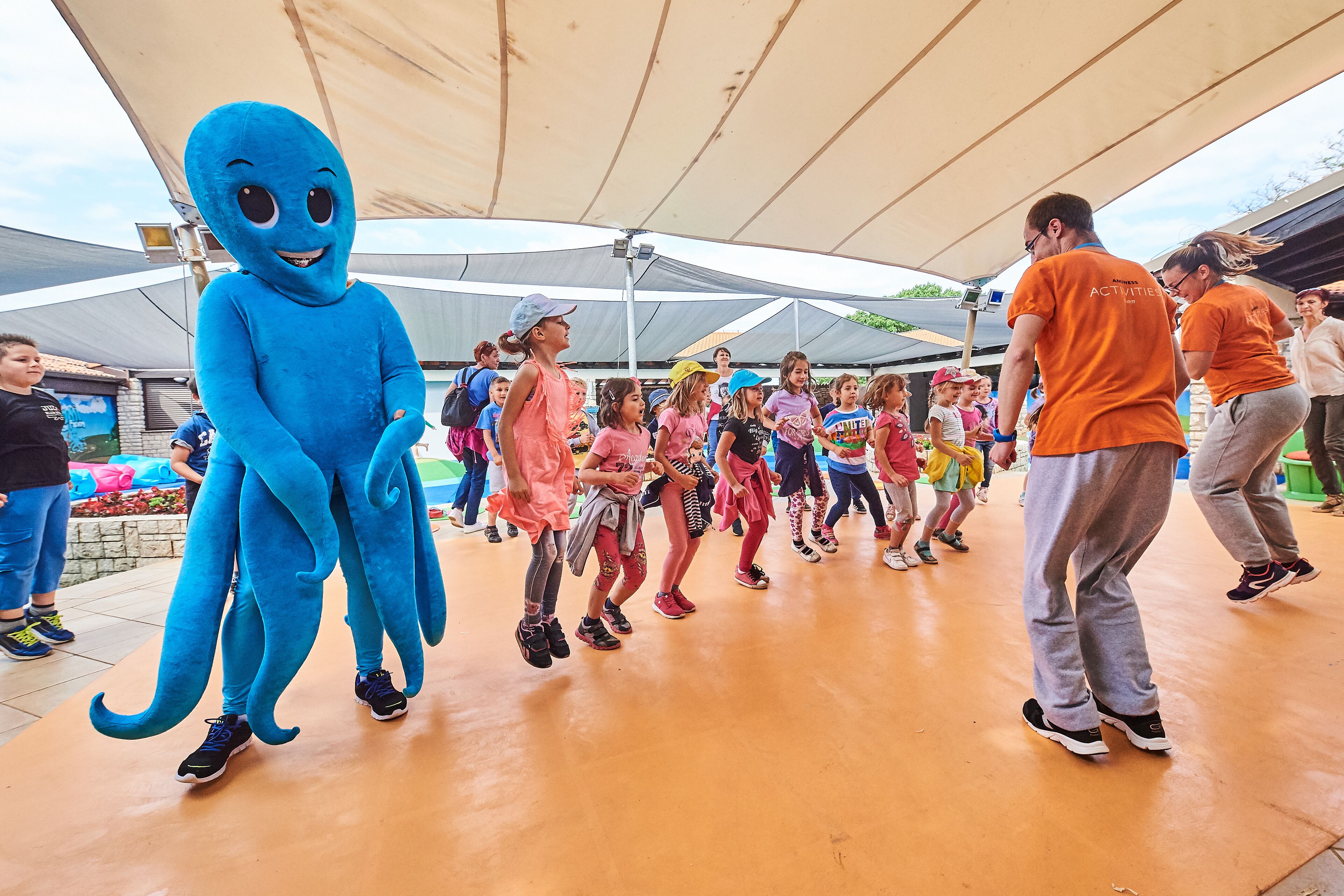 A group of children in a line under a tent, with a person in a blue costume standing to the side.