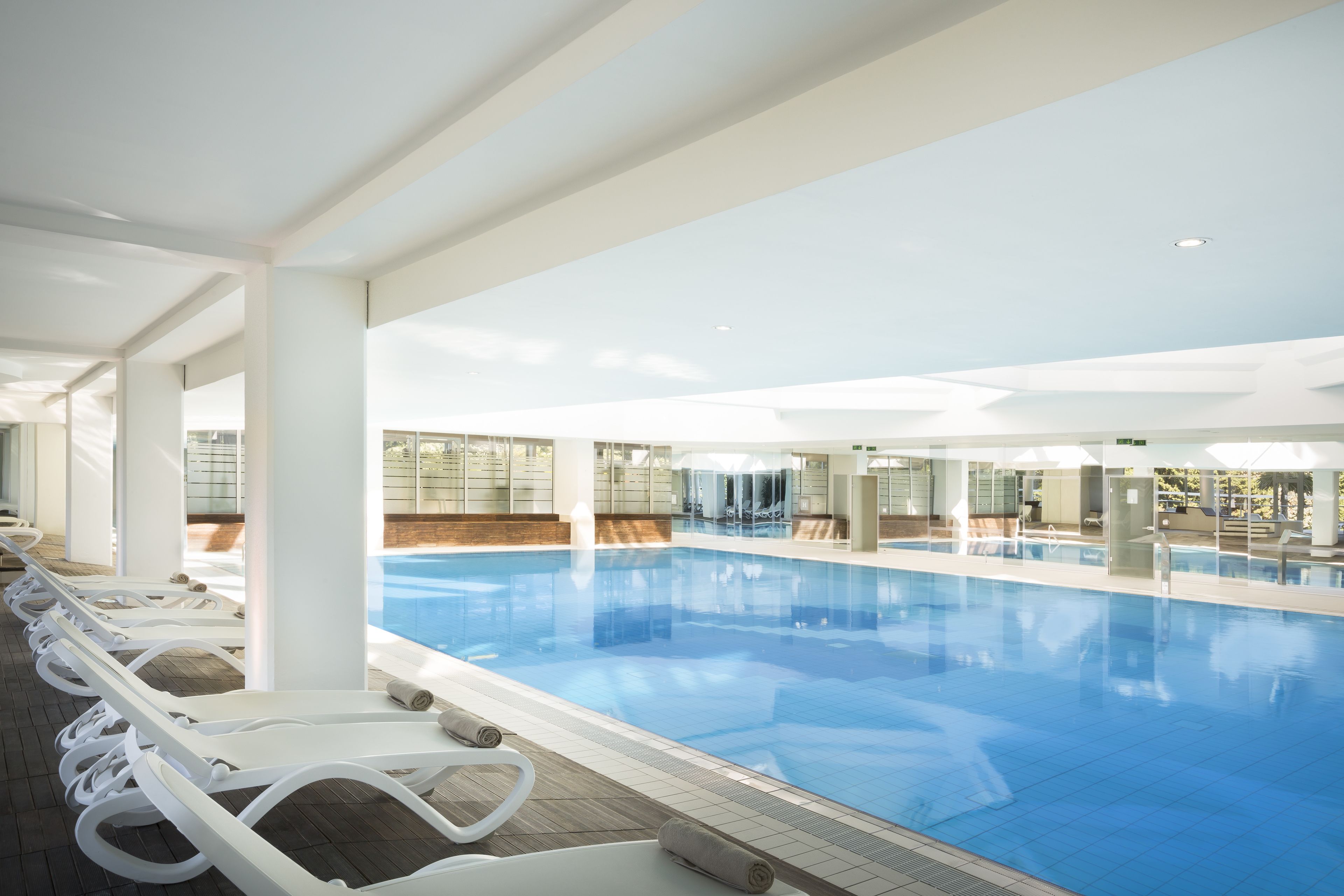 Una piscina interior moderna con agua azul cristalina, rodeada de tumbonas blancas y suelo de baldosas.