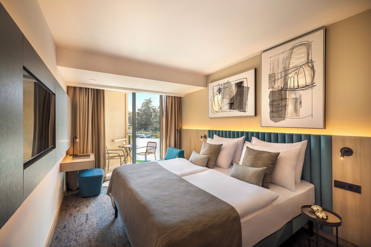 Habitación de hotel con una cama grande, un escritorio y una silla. Hay dos ventanas amplias y vistas al exterior.