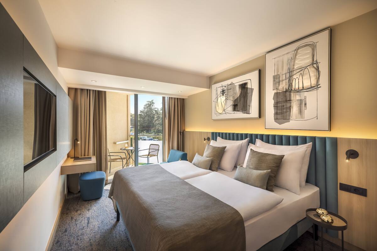 A hotel room with a large bed, a desk, and a chair. There are two large windows with curtains, a view of the outside, and artwork on the wall.