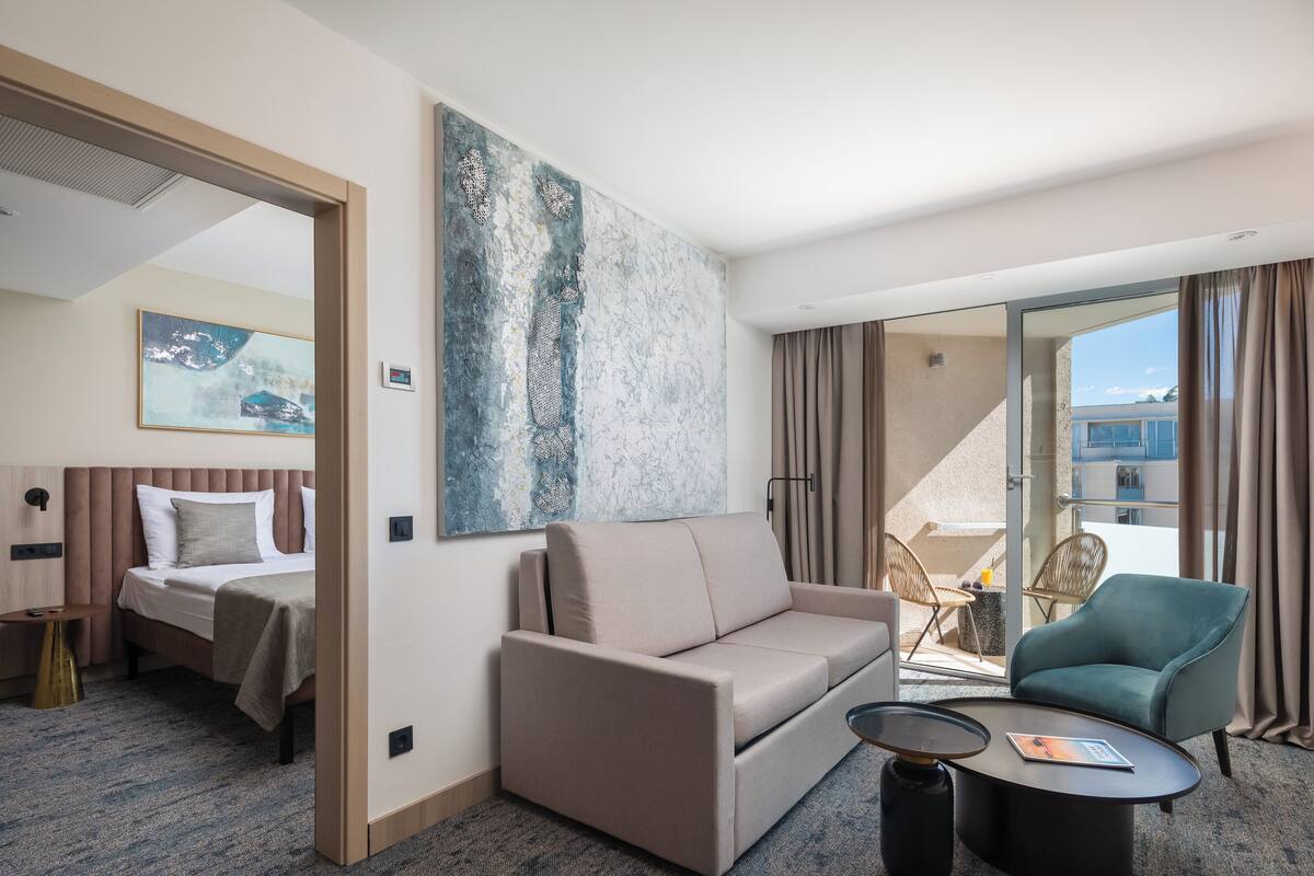 A modern hotel room with a bed, sofa, and armchair, a large abstract painting, and a balcony with curtains.