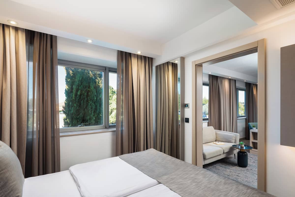 A modern hotel room with a large bed, a sofa, a small table, and a view of greenery outside.