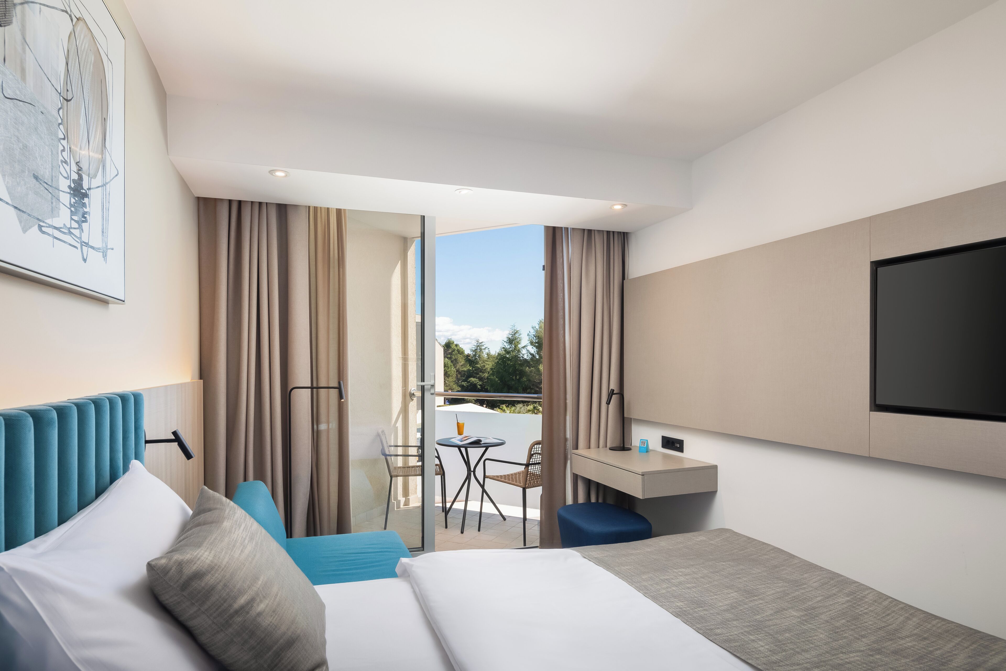 A modern hotel room with a bed, a desk, a TV, and a balcony with a table and chairs.