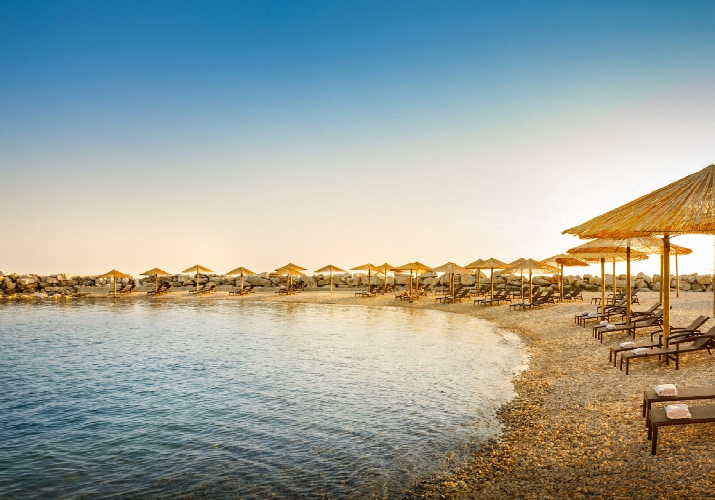 A beach with rows of sun loungers and umbrellas, a calm sea, and a rocky shoreline.