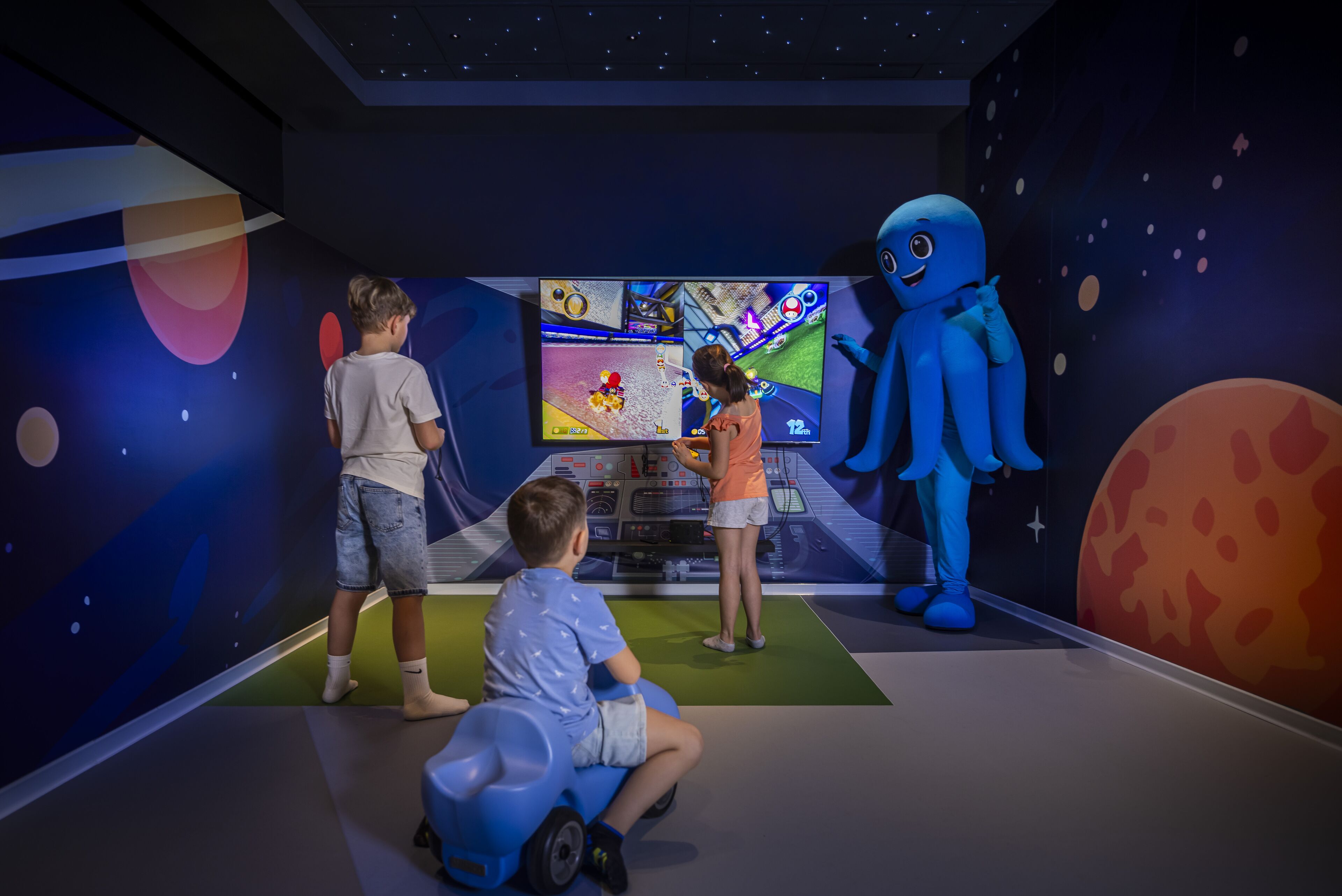 Een kamer met een groot scherm waarop een spel wordt getoond, een kind op een kinderauto en een mascotte.