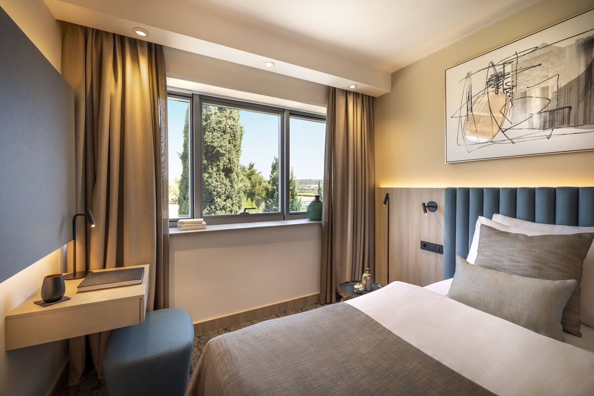 A hotel room with a large bed, a desk with a chair, a window with curtains, a wall-mounted artwork, and a nightstand with a lamp.