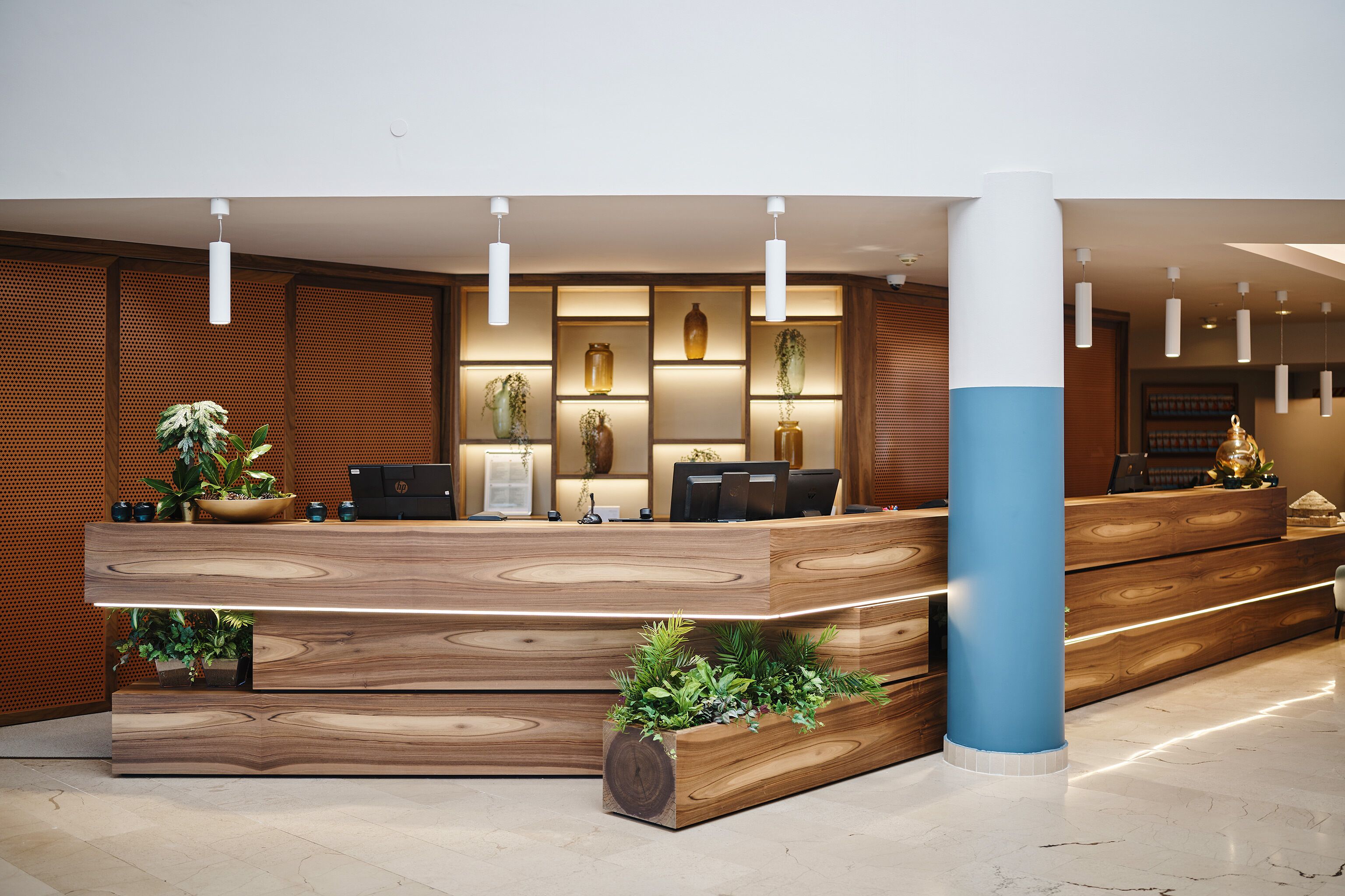 A reception desk with a wooden finish, integrated plants, and a blue pillar.