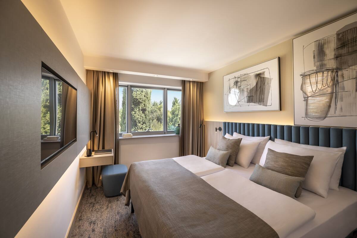 A hotel room with a large bed, a desk, a chair, a window with curtains, and artwork on the walls.