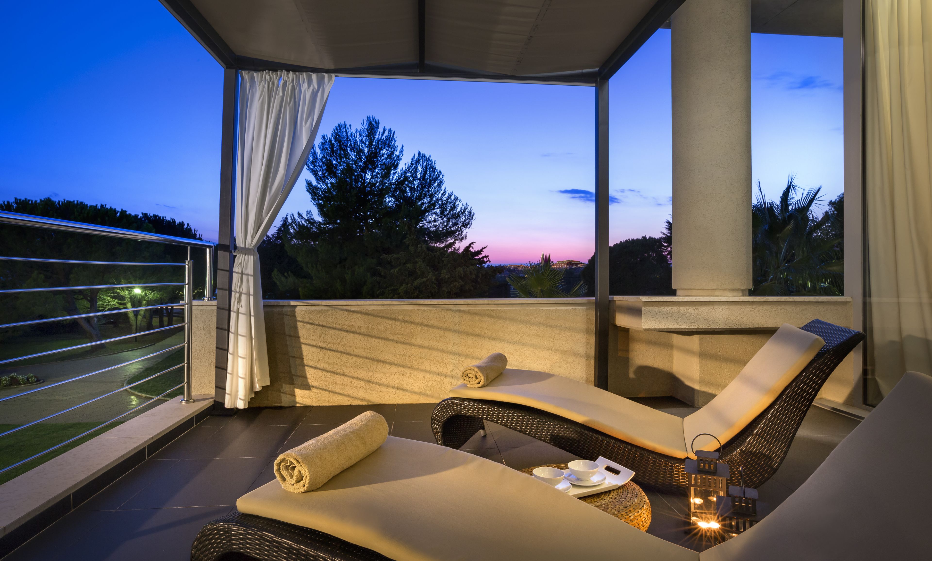 A balcony with two lounge chairs, a table with a bowl and a cup, and a view of trees and a sunset.