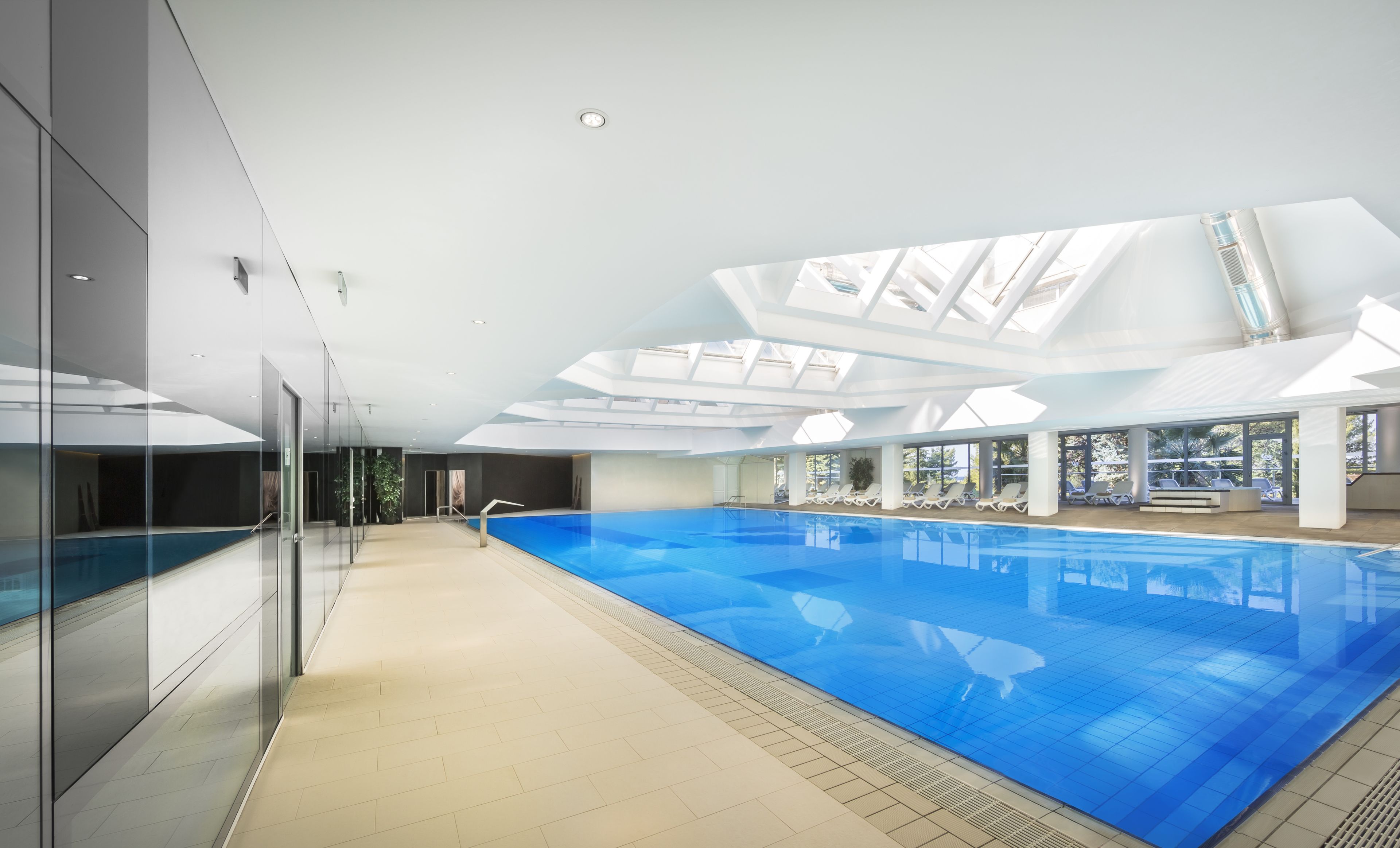 Une piscine intérieure moderne, dotée d’une structure en verre, de lucarnes et d’un espace détente baigné de lumière naturelle.