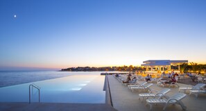 Une piscine à débordement offrant une vue sur la mer, des chaises longues et un bâtiment abritant un bar.