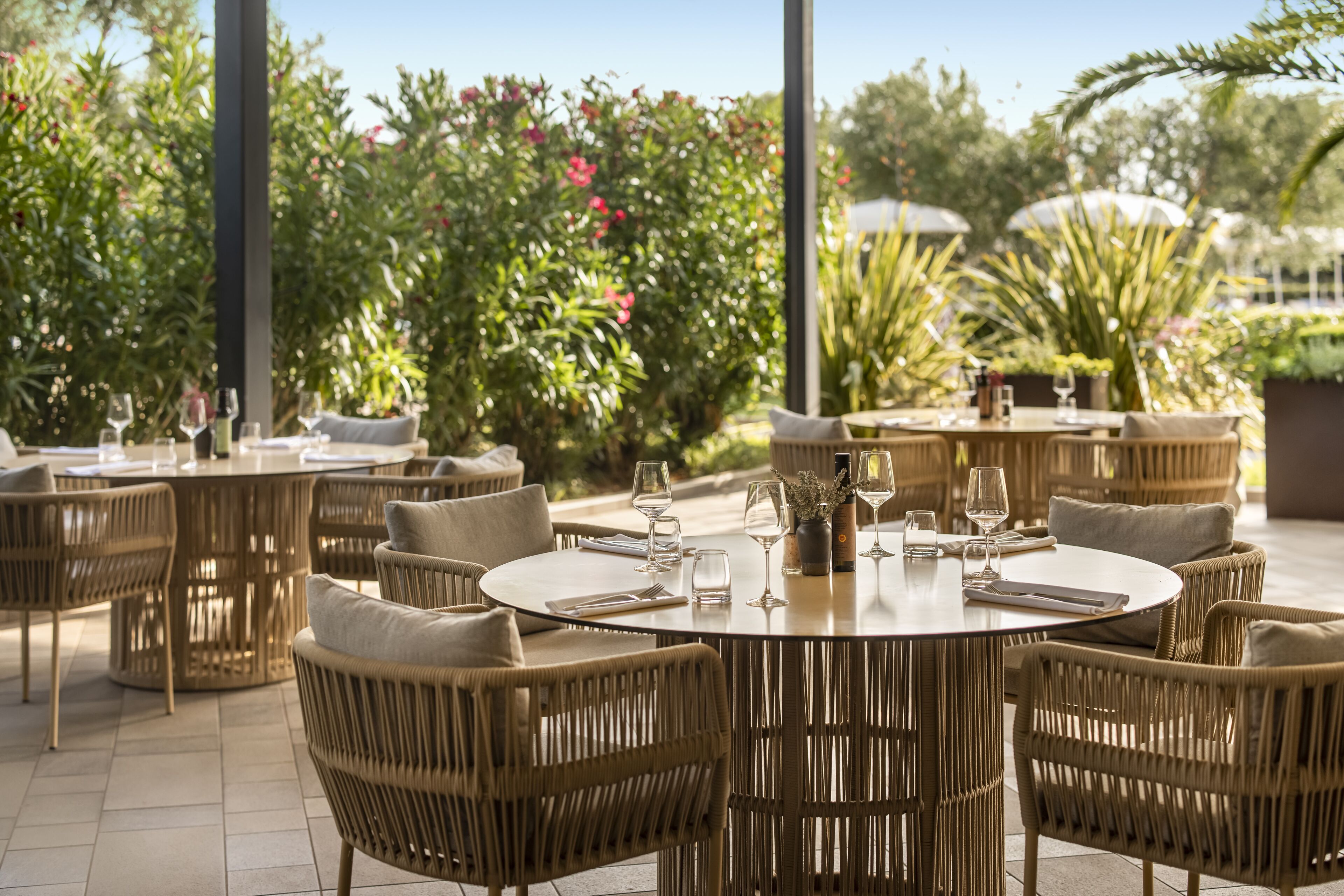 Un área de comedor al aire libre con mobiliario de mimbre y mesas redondas, rodeada de abundante follaje y plantas.