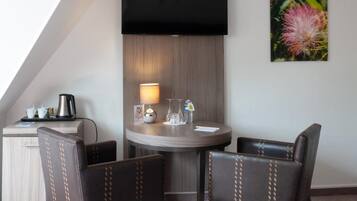 A hotel room with a TV mounted on the wall, a round table with two chairs, a small desk with a coffee maker, and a lamp.