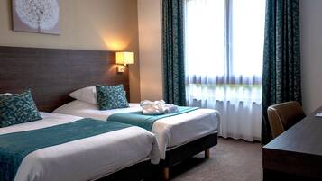A hotel room with two beds, a desk, a lamp, and a window with curtains.