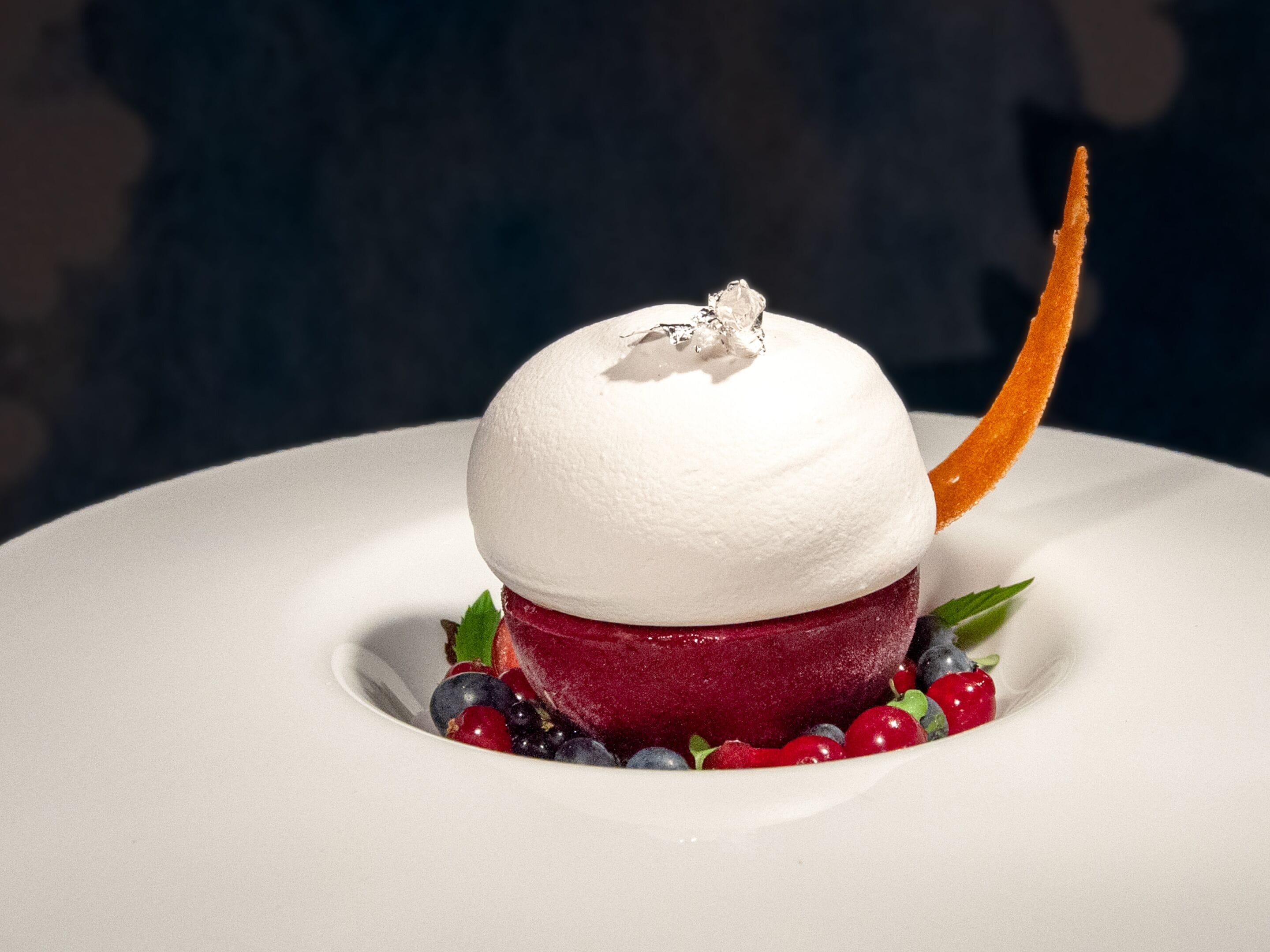 A dessert with a white dome, red base, and berries, garnished with a leaf and a thin orange slice.