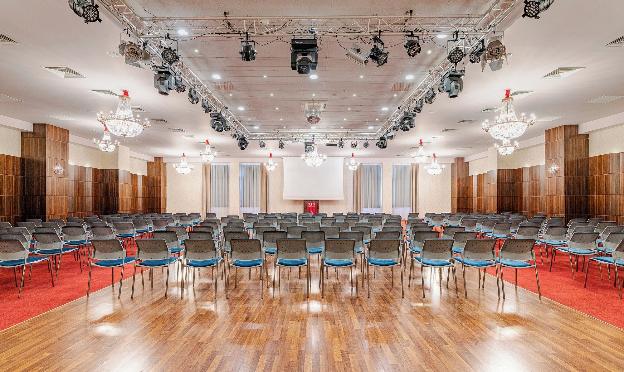 Een vergaderzaal met rijen stoelen, een groot scherm en verlichtingsarmaturen aan het plafond.