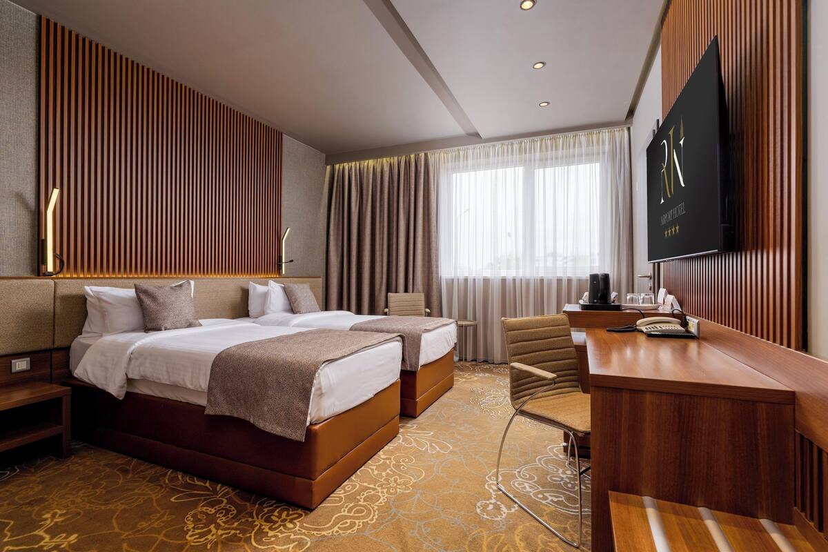 A hotel room with two beds, a desk, a TV, and a large window with curtains.
