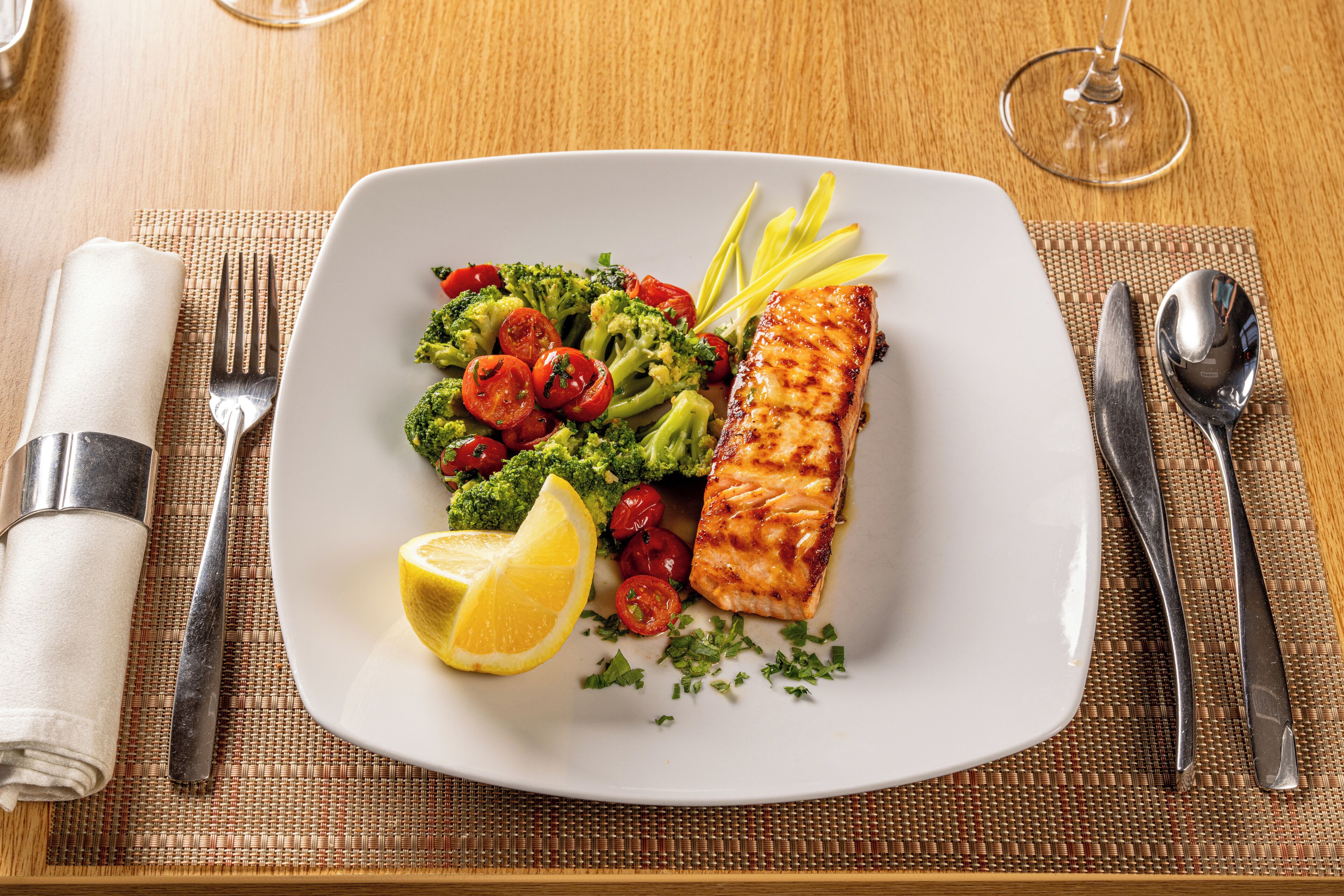Een witte schaal met gegrilde zalm, broccoli, kerstomaatjes en een citroen op een houten tafel.