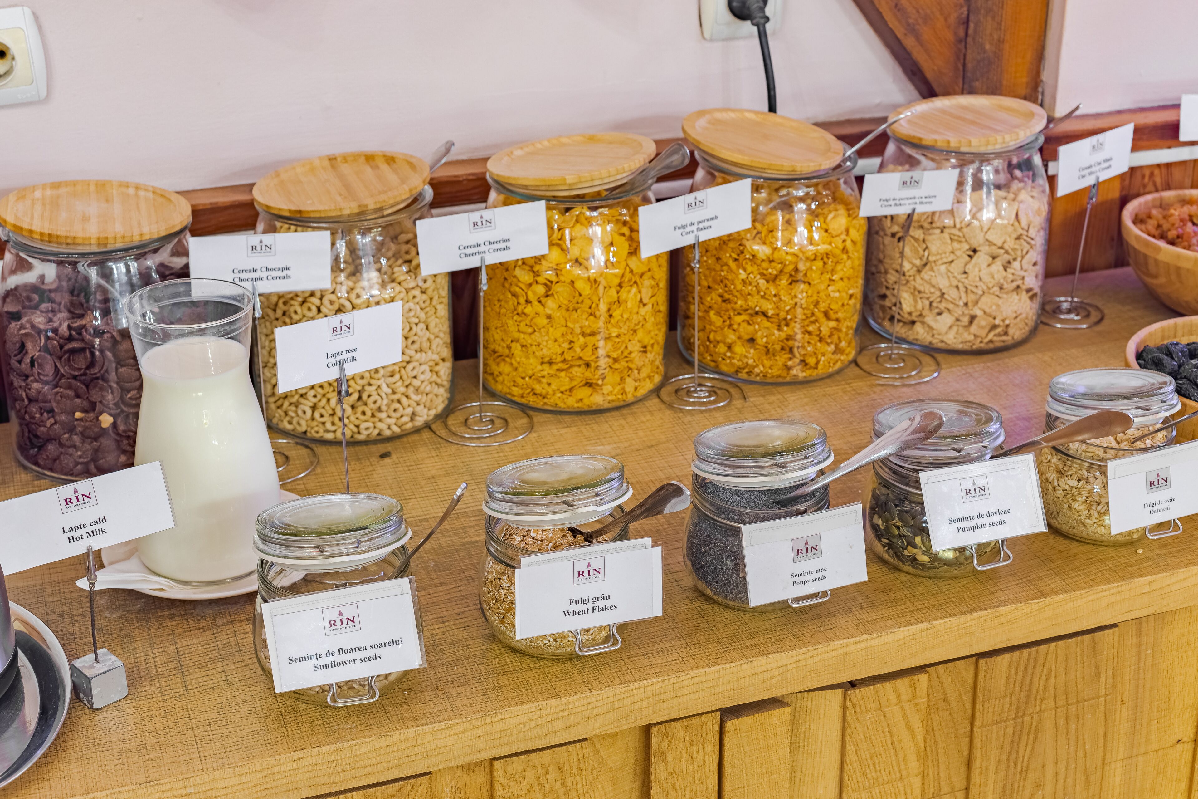 Diverse levensmiddelen in glazen potjes met etiketten, waaronder ontbijtgranen, zaden en melk.
