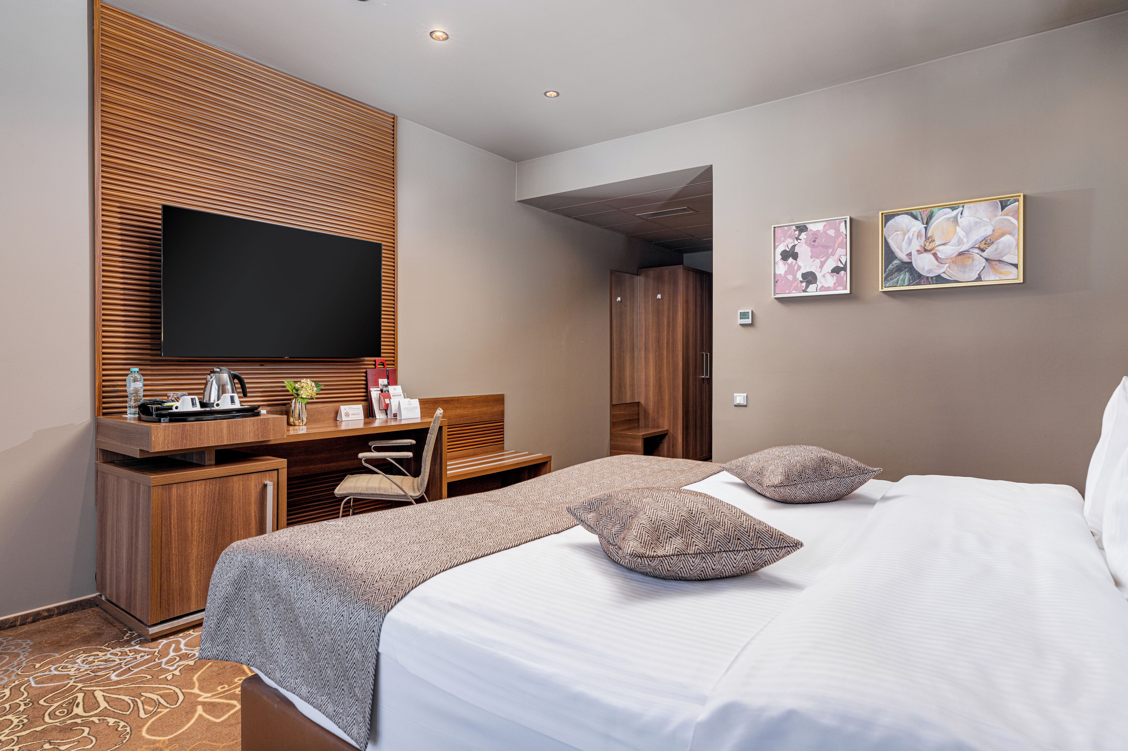 A modern hotel room with a large bed, a flat-screen TV mounted on a wooden panel, a desk with a chair, and two framed floral artworks on the wall.