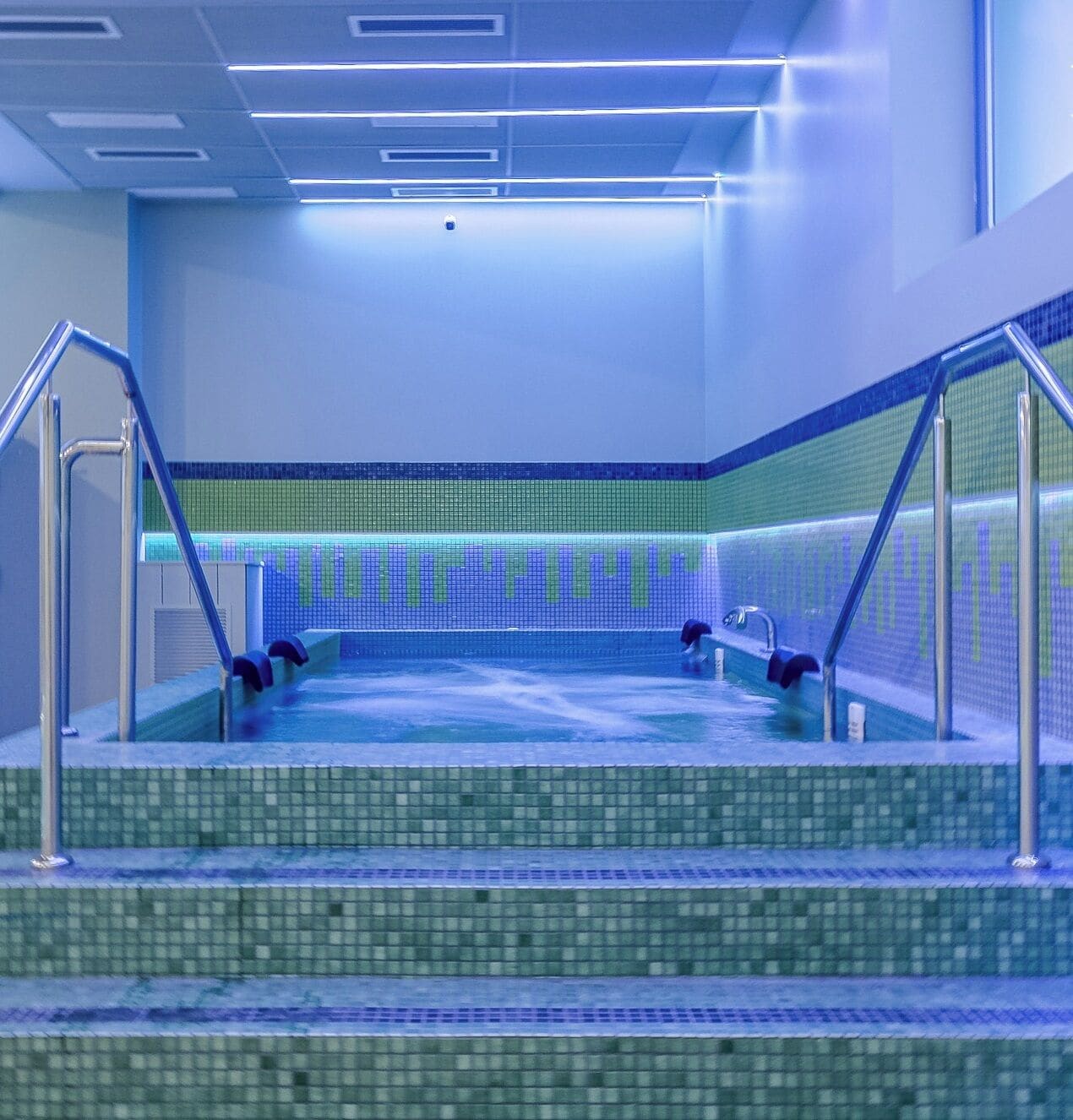 A modern indoor swimming pool with clear water, tiled walls, and metal railings.