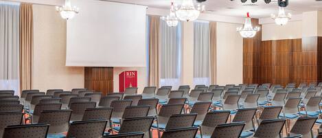 A conference room with rows of chairs, a large screen, and chandeliers.