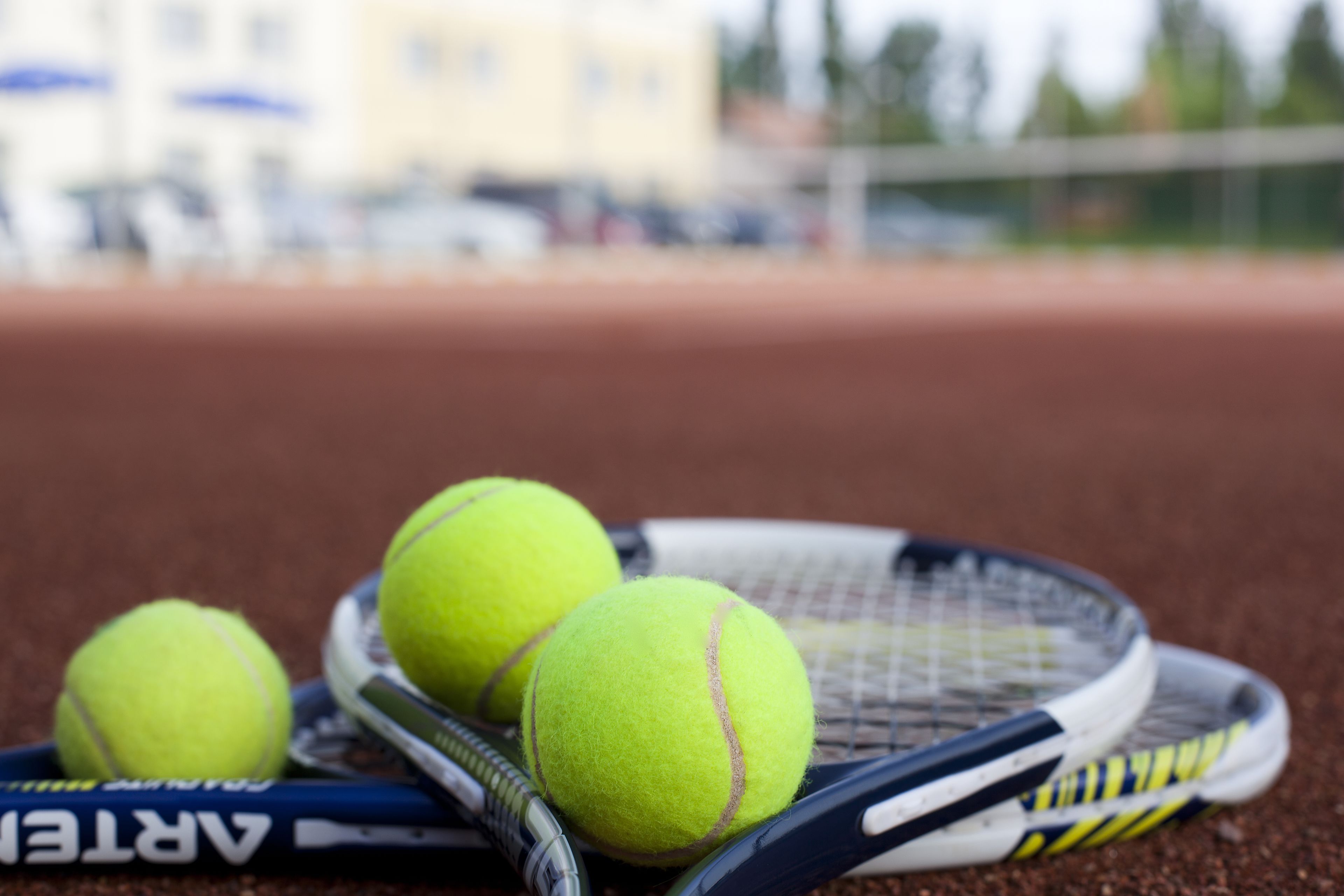 A tennis racket and three tennis balls on a clay court.