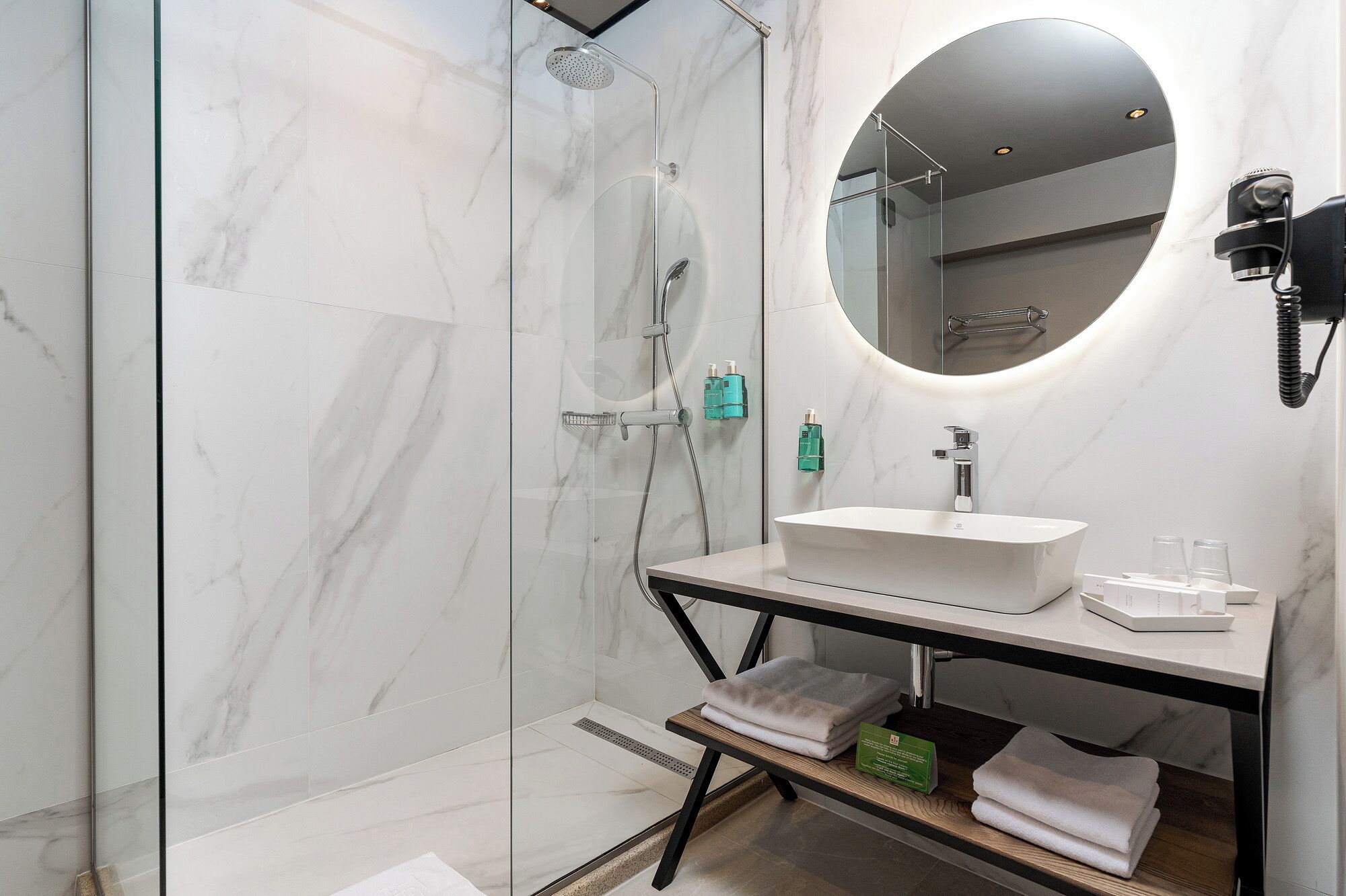 A modern bathroom with a glass-enclosed shower, a white sink, and a round mirror.