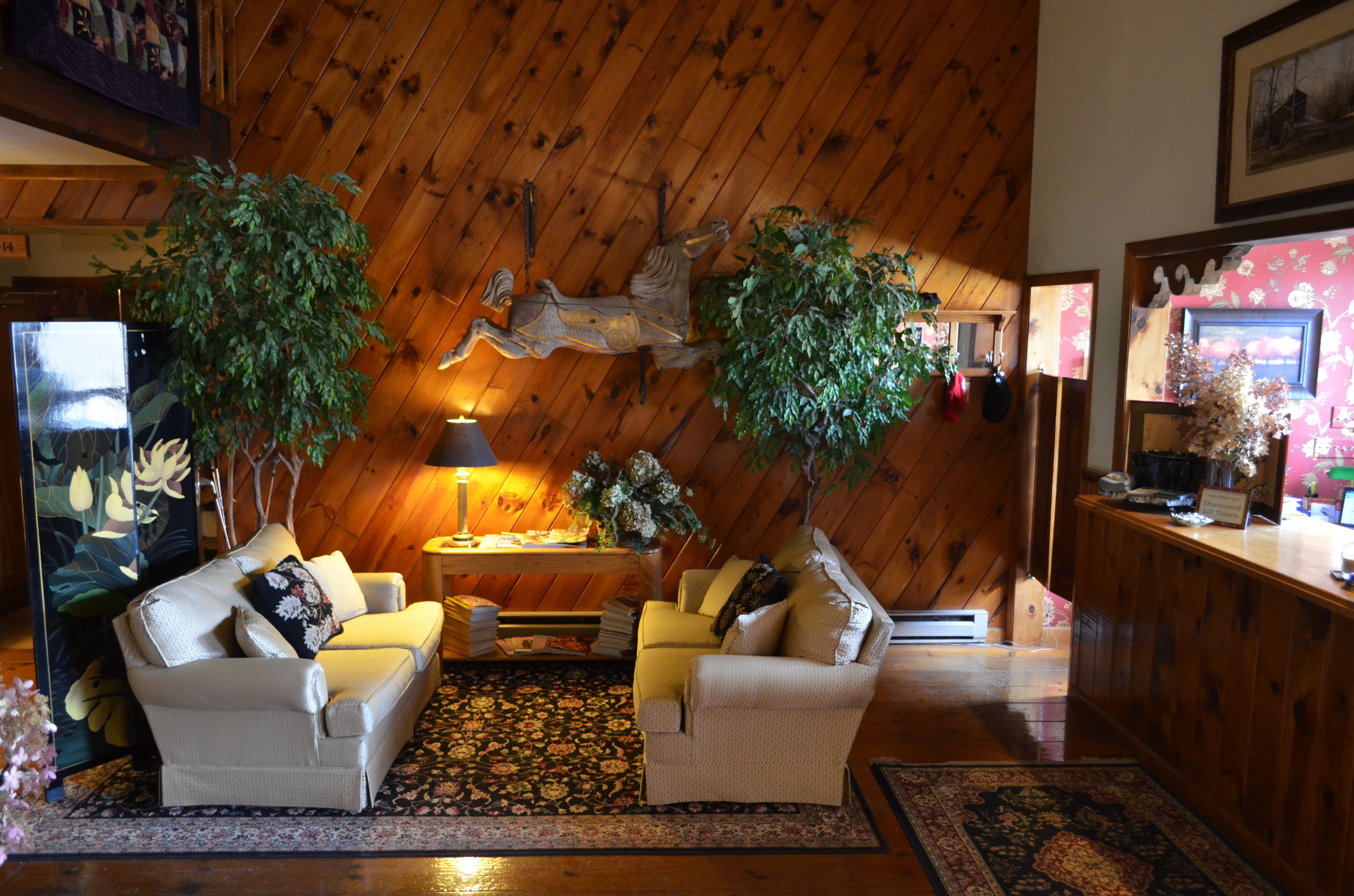 A cozy living room with two sofas, a coffee table, and a fireplace. There are two large potted plants and a wooden wall with a horse decoration.