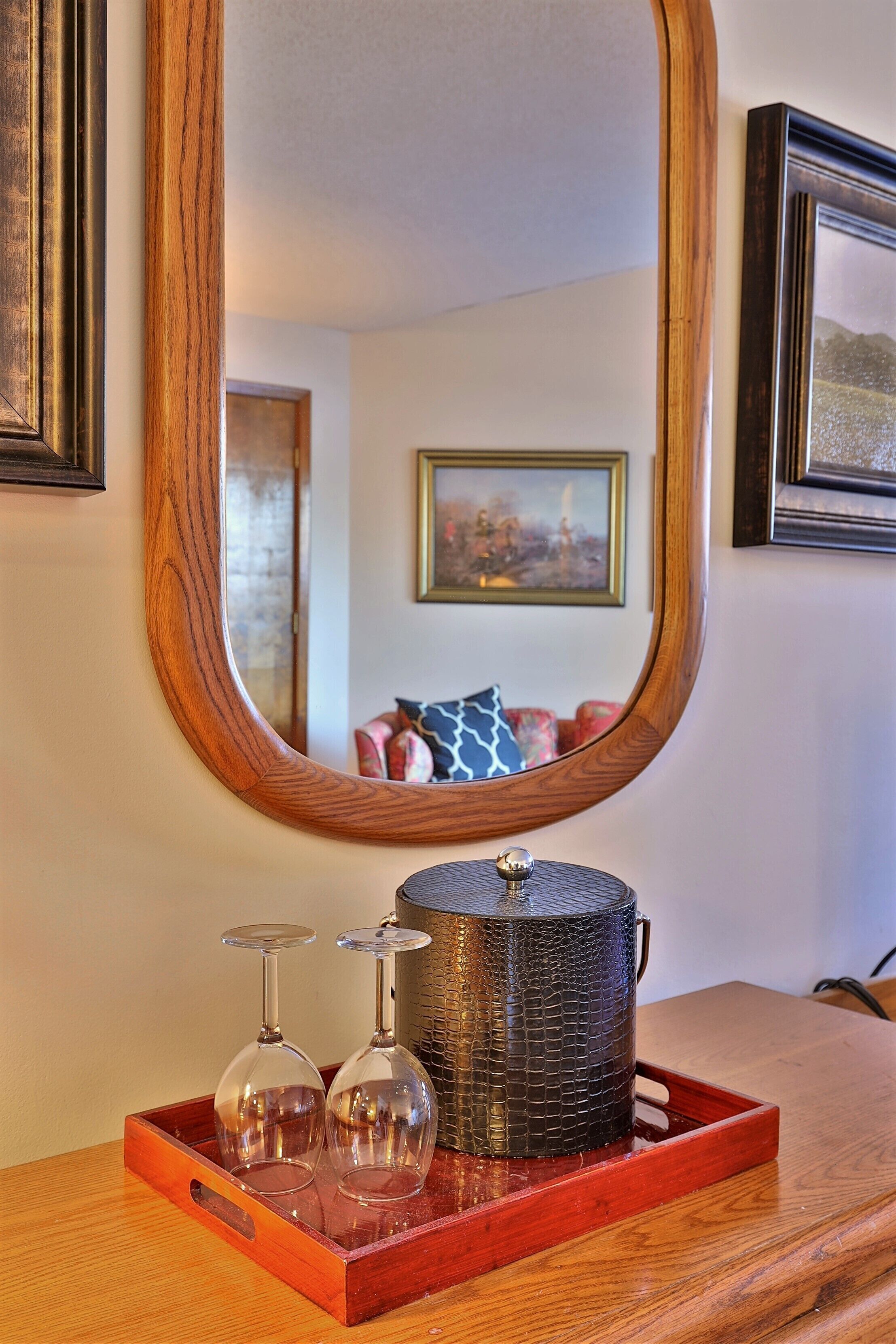 Una bandeja de madera con dos copas de vino y un recipiente decorativo sobre una mesa de madera, reflejando un cuadro enmarcado y un sofá con cojines estampados al fondo.