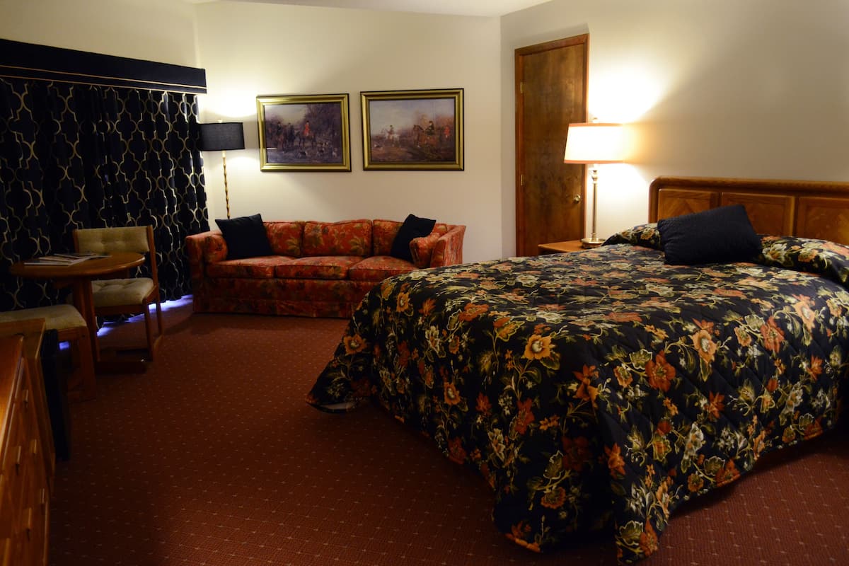 A hotel room with a bed, a sofa, a chair, a lamp, and two framed pictures on the wall.