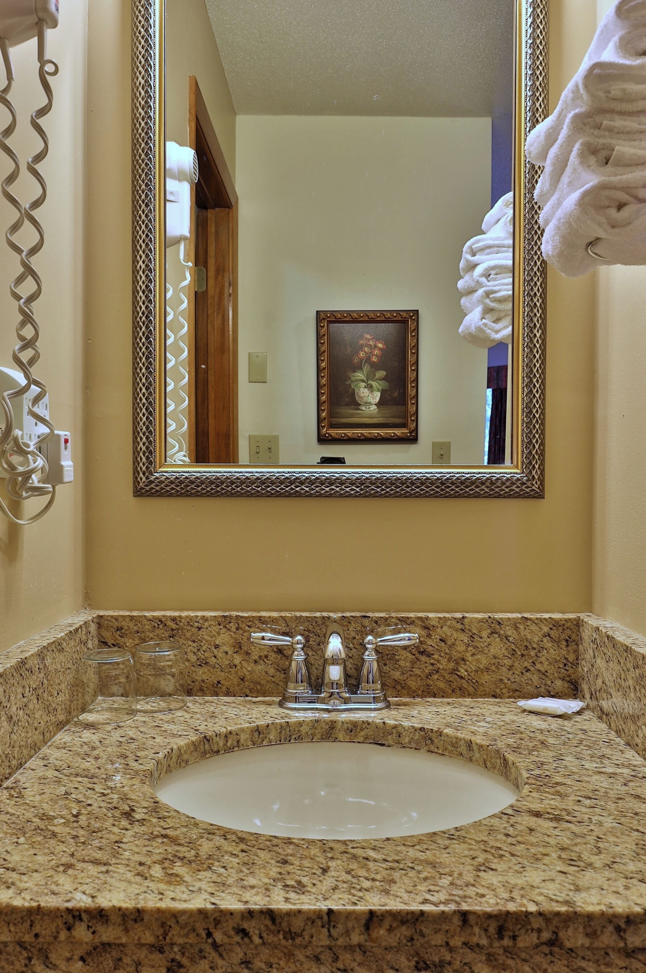 Une salle de bain dotée d’un lavabo avec un plan de travail en granit, d’un miroir orné d’un cadre décoratif et d’un porte-serviettes sur lequel sont pliées des serviettes.
