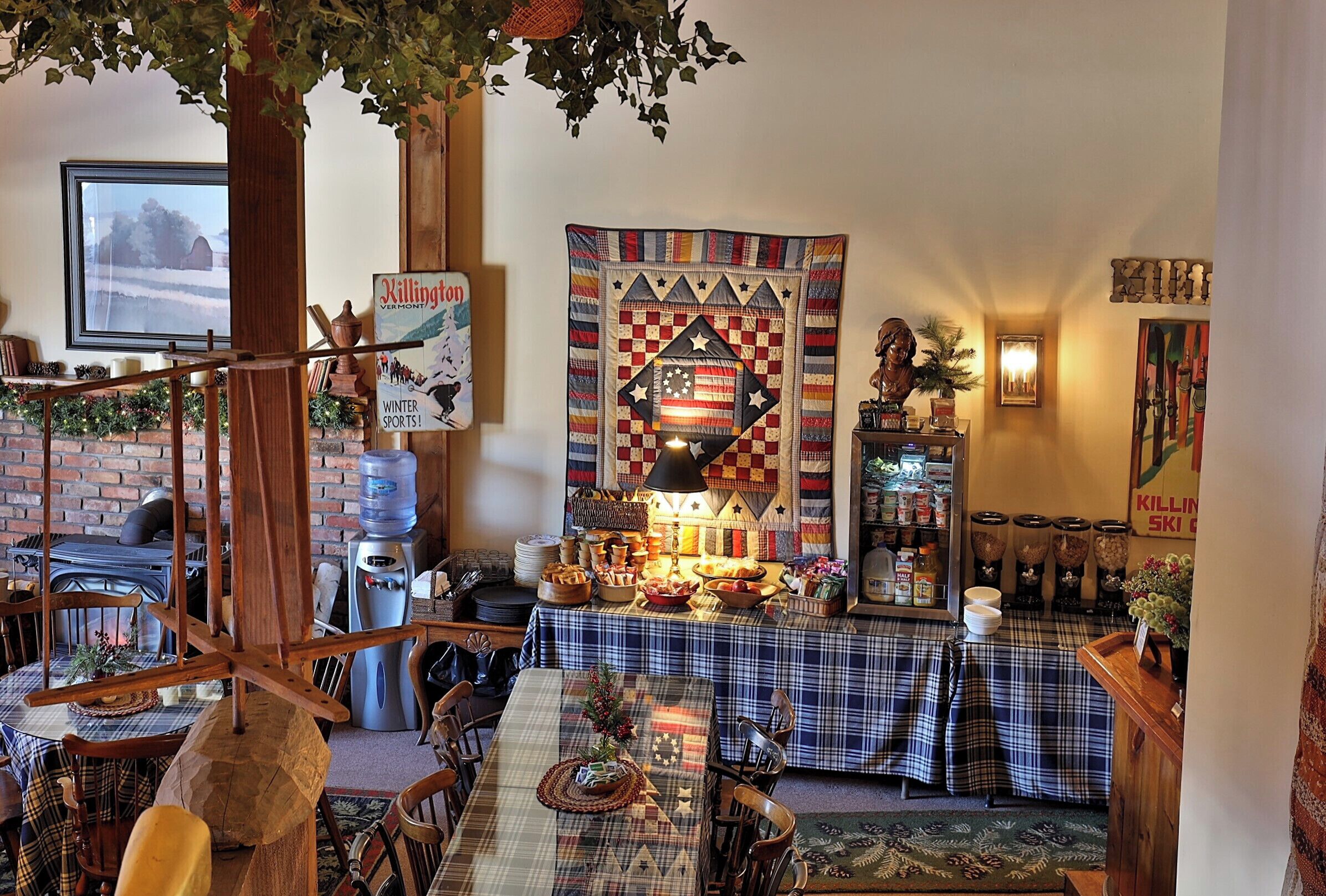 A cozy dining area with a large table set for a meal, surrounded by chairs. A wooden staircase is visible in the foreground, and a large quilt hangs on the wall.