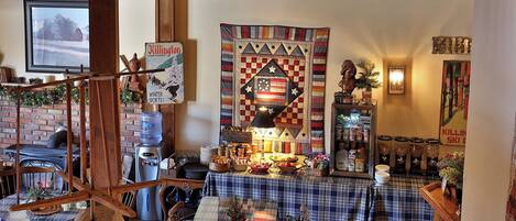 A cozy dining area with a large table set for a meal, surrounded by chairs. A wooden staircase is visible in the foreground, and a large quilt hangs on the wall.
