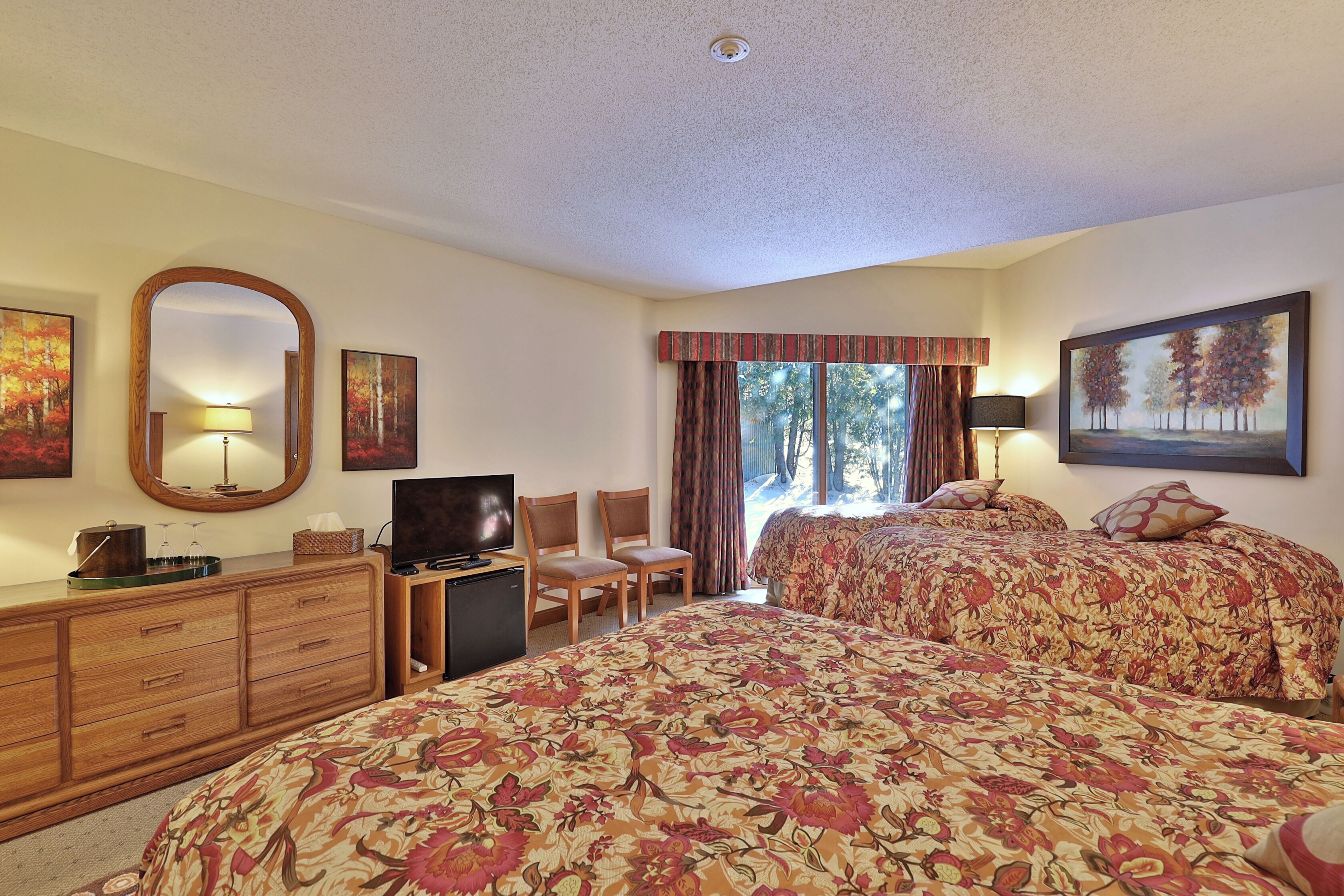 A hotel room with two beds, a TV, a desk with a lamp, and a dresser.