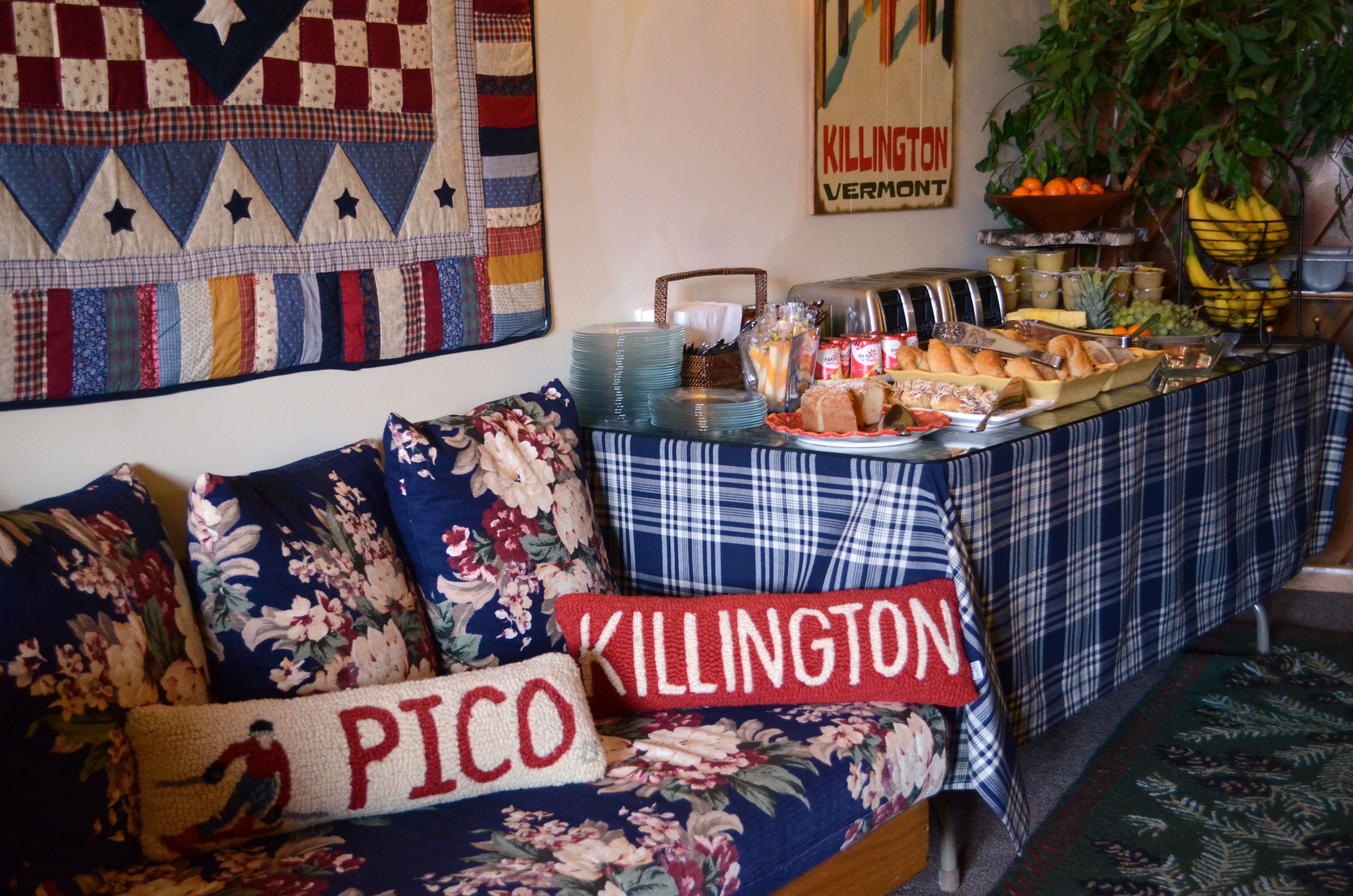 A room with a quilted wall hanging, a table with breakfast items, and a couch with floral pillows.