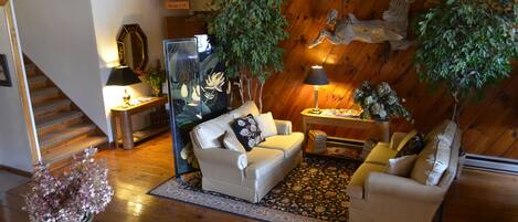 Uma sala de estar aconchegante com sofá, poltronas e uma mesa de centro. Há duas plantas em vasos, uma lareira e uma escadaria que leva a outro cômodo.
