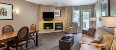 A hotel room with a dining table, chairs, a sofa, a TV, a fireplace, and a lamp.