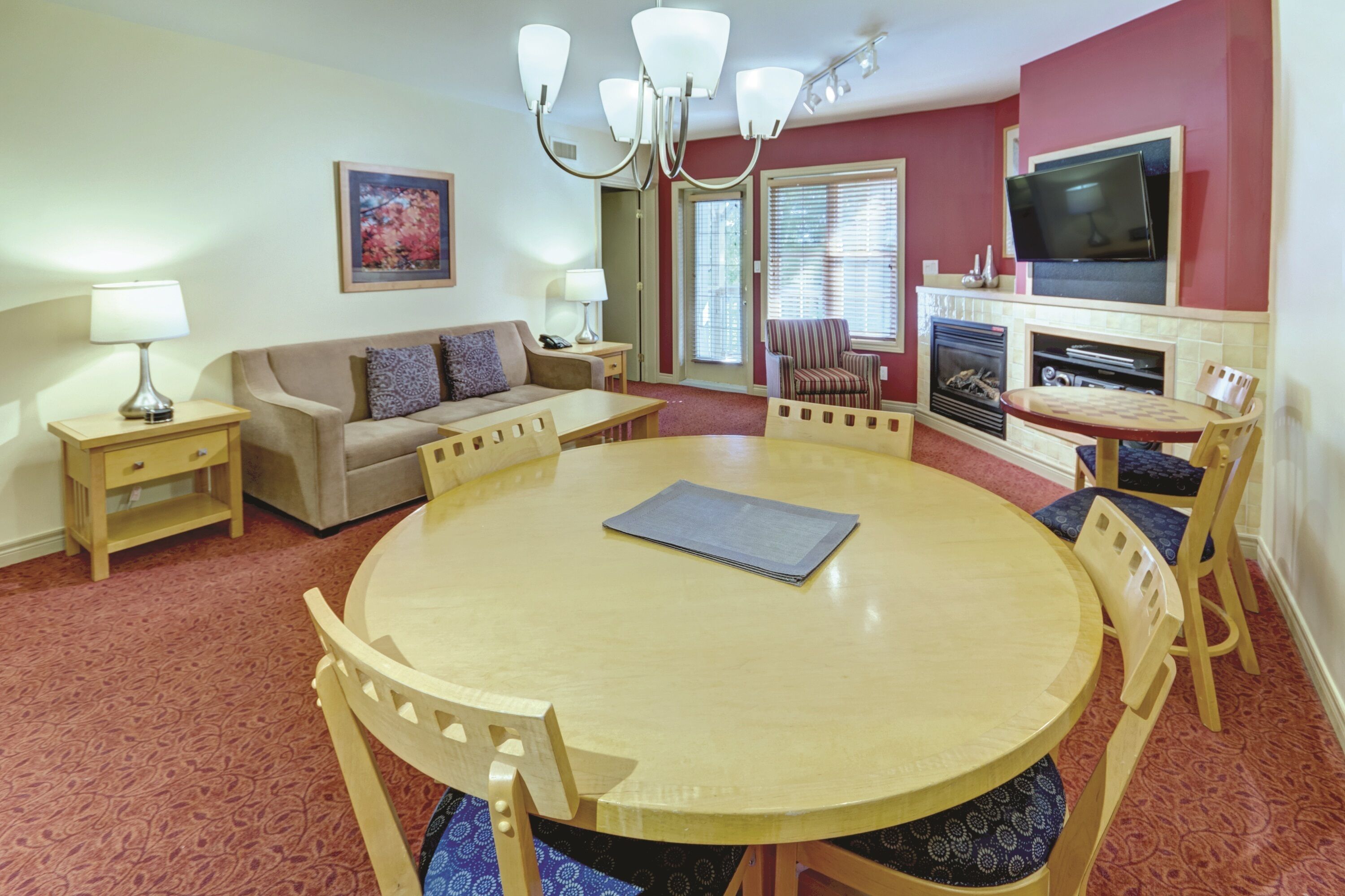 A living room with a round wooden table, beige sofa, striped armchair, and a flat-screen TV mounted on the wall.