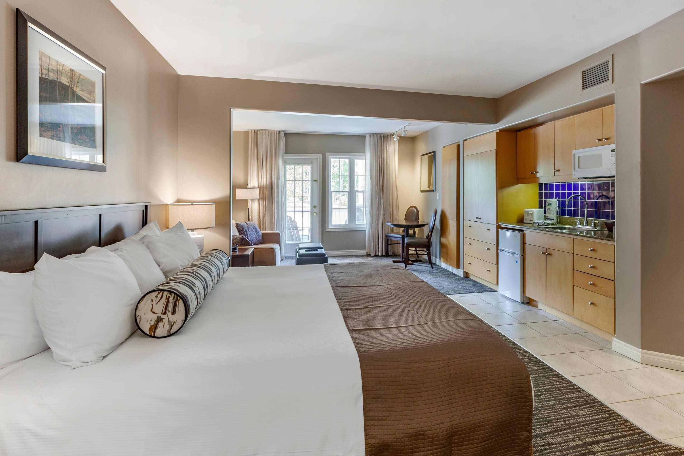 A hotel room with a large bed, a desk, a chair, and a kitchenette.