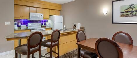 A modern kitchen with wooden cabinets, a white refrigerator, a microwave, and a coffee maker. There is a dining table with four chairs and a rug on the floor.