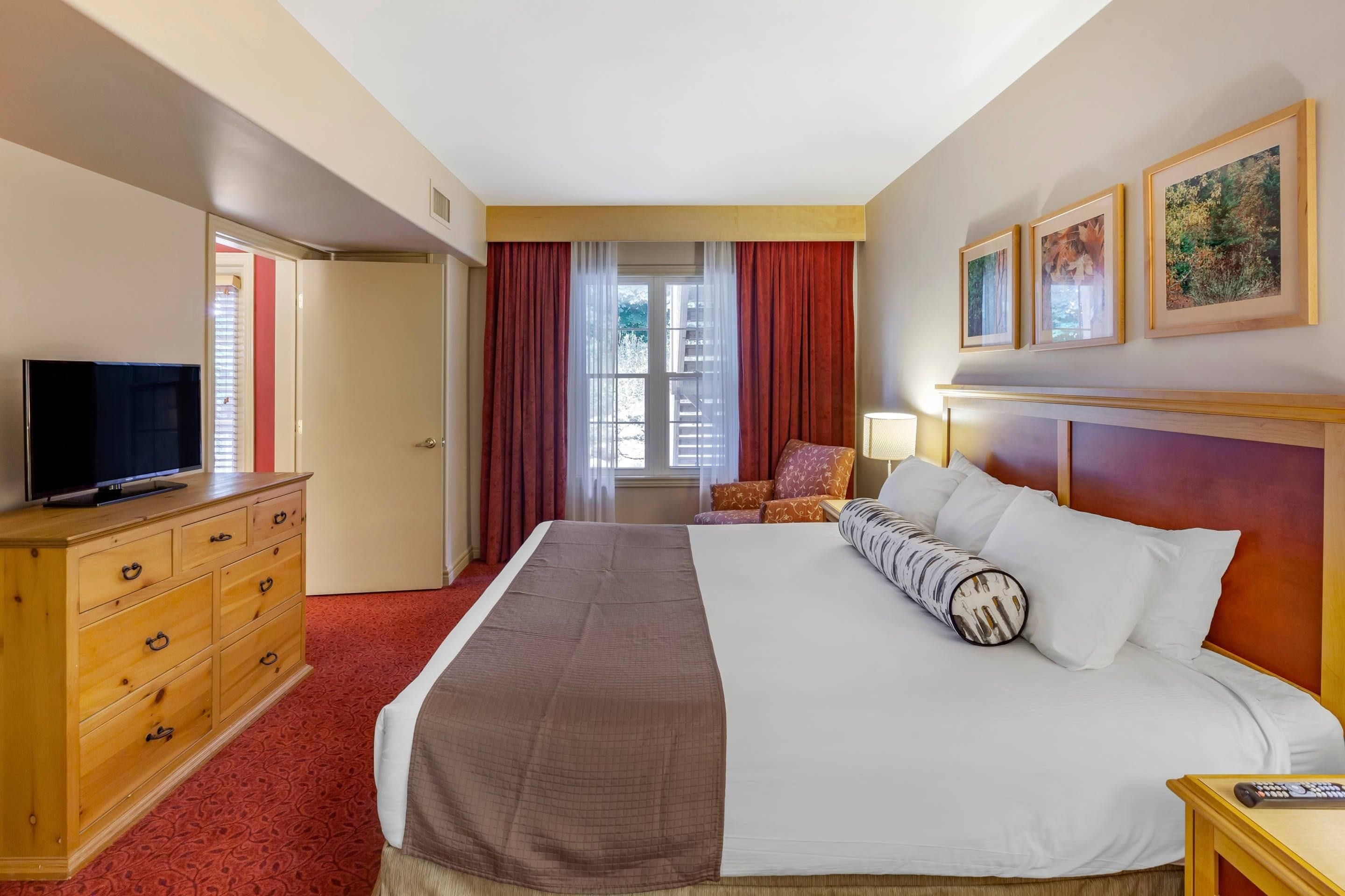Une chambre d’hôtel avec un grand lit, une commode en bois, une télévision, une chaise et une fenêtre avec des rideaux.
