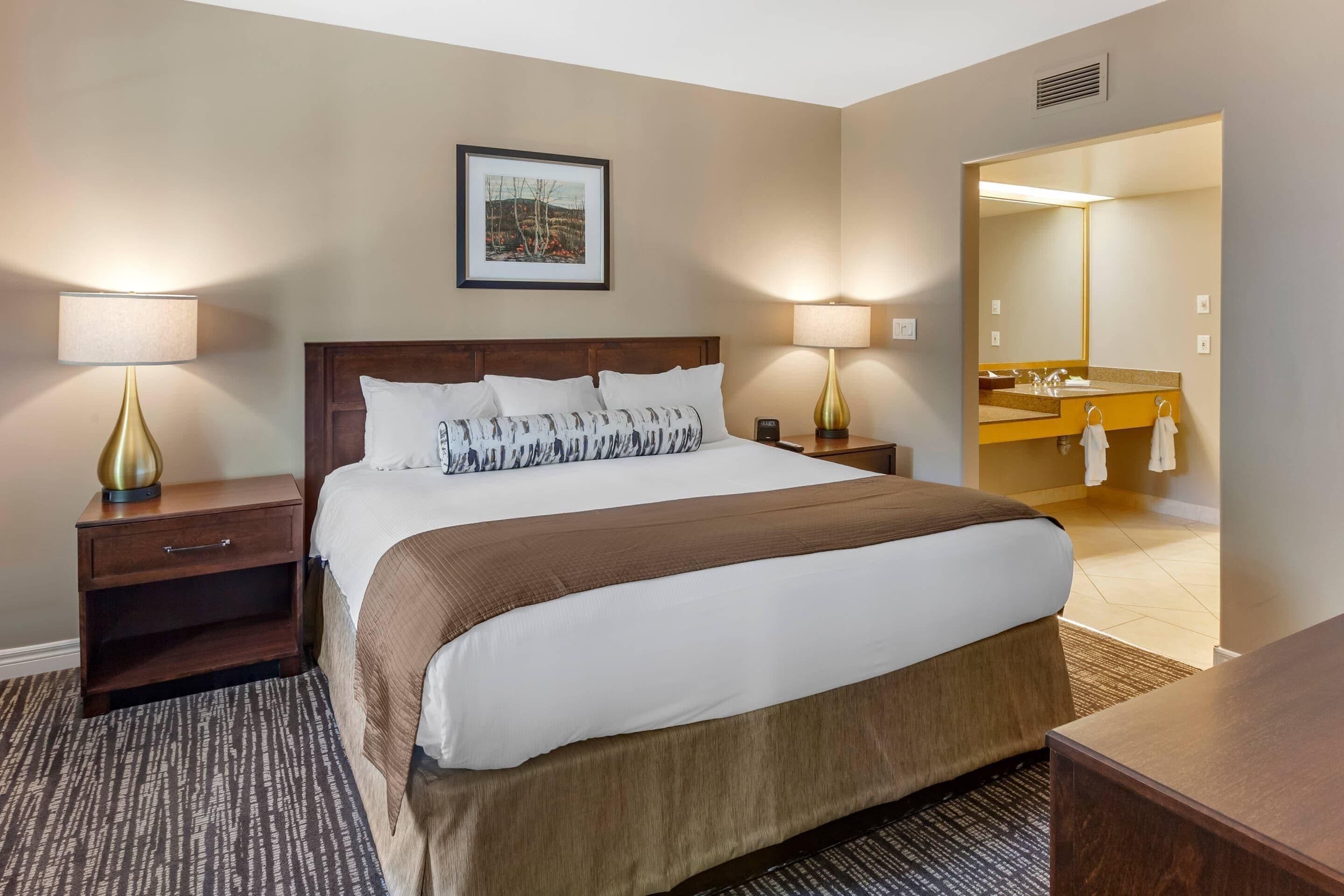 Une chambre d’hôtel avec un grand lit, deux tables de chevet avec des lampes, un tableau encadré au mur et une salle de bain visible à travers une porte ouverte.