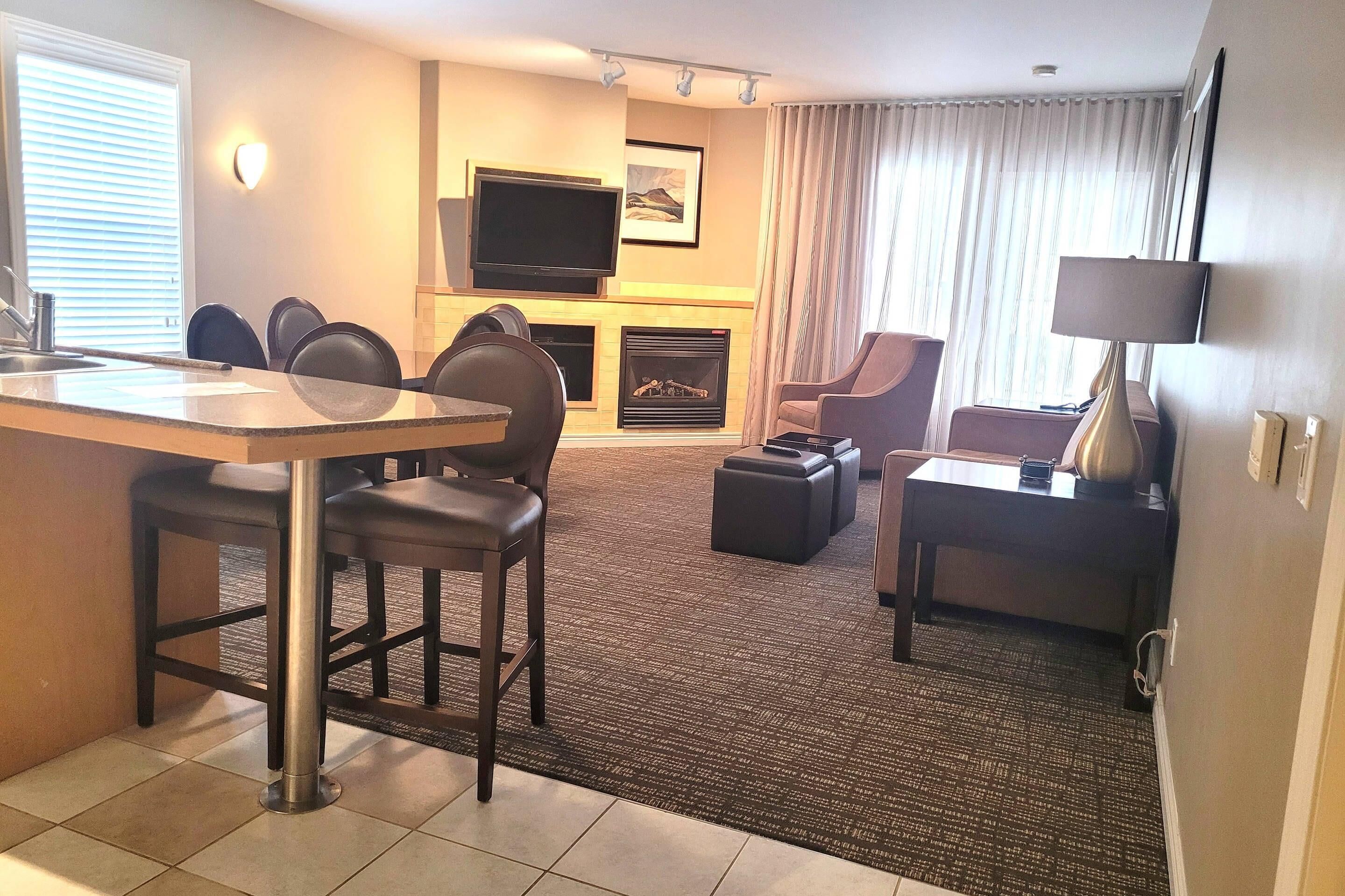 A hotel room with a dining area, a flat-screen TV mounted above a fireplace, a sofa, and a lamp.