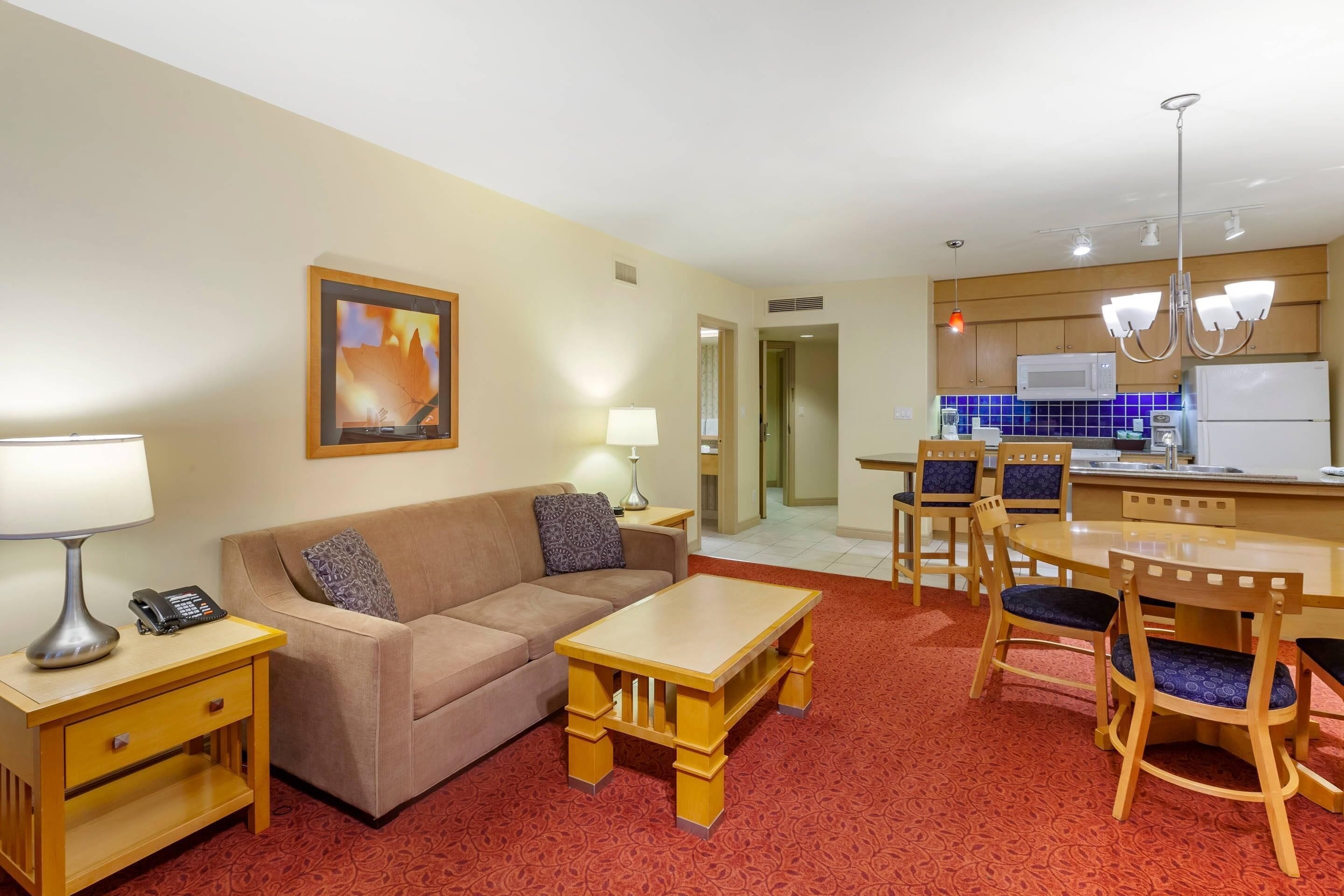 A hotel room with a sofa, a coffee table, a dining table with chairs, a kitchen area with a microwave, and a refrigerator.