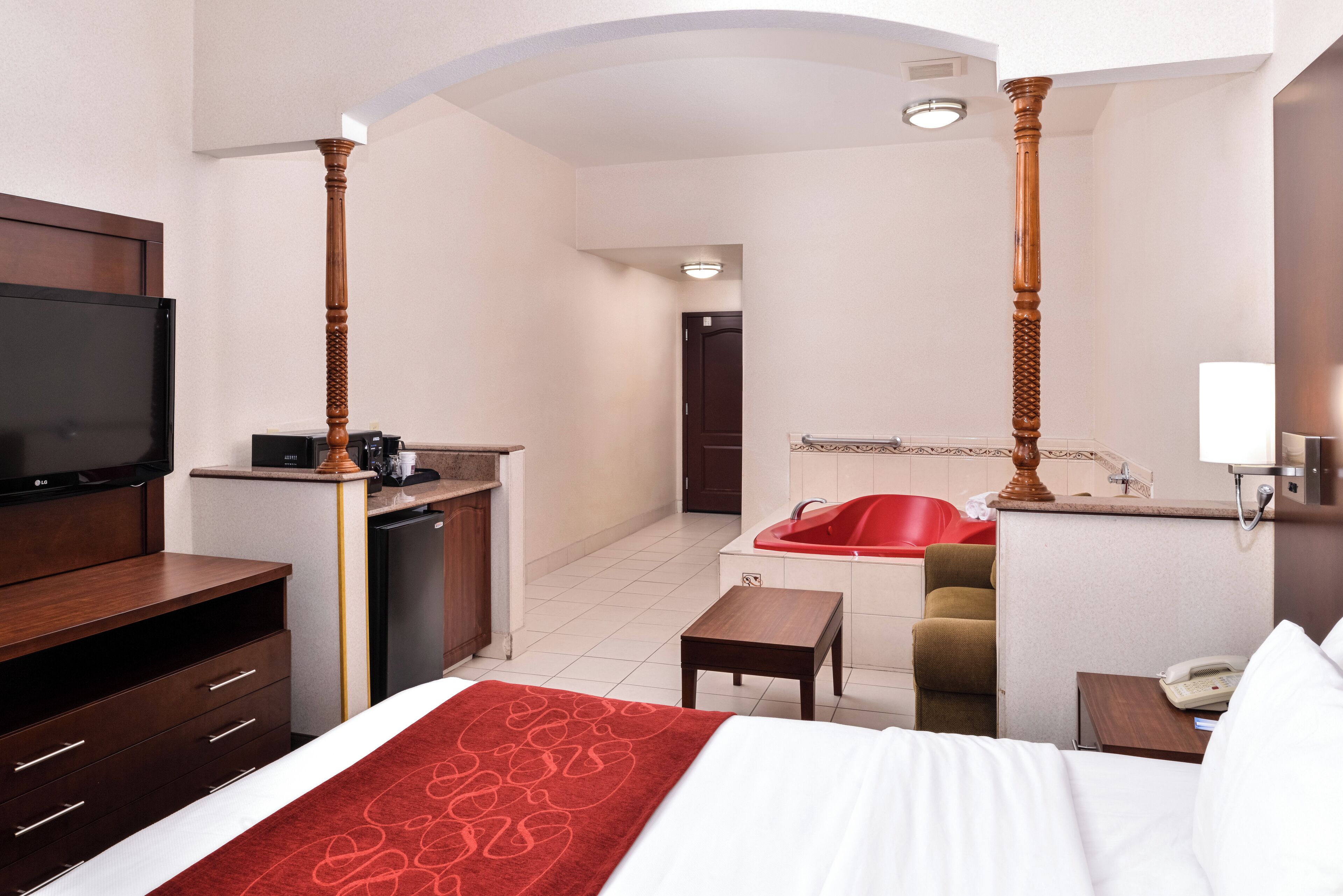 A hotel room with a bed, a TV, a desk with a computer, a red bathtub, and a small table.