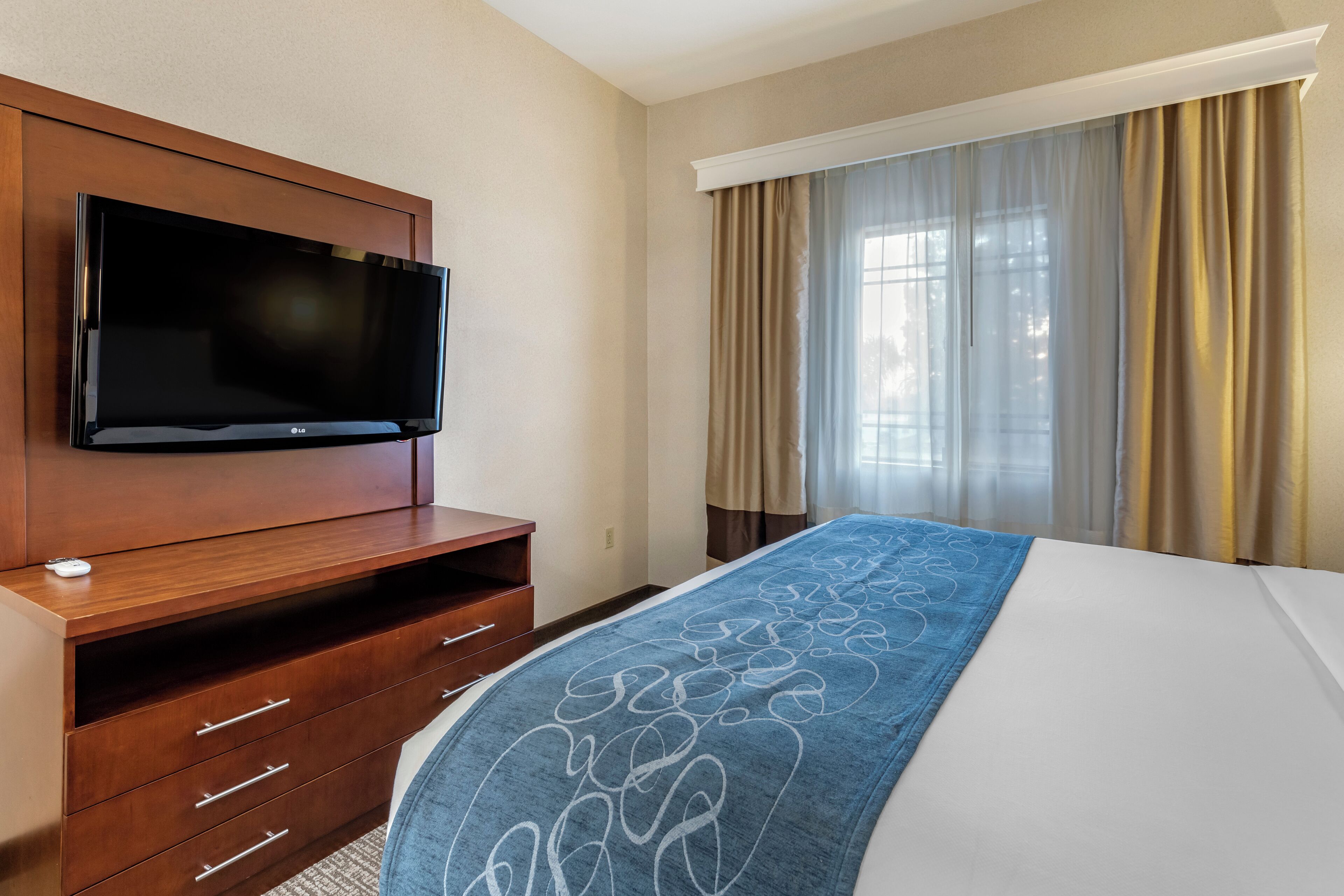 Une chambre d’hôtel avec un téléviseur à écran plat fixé sur un meuble en bois, un lit recouvert d’une couverture bleue et une fenêtre avec des rideaux beiges.