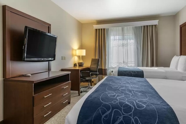 Habitación de hotel con televisor de pantalla plana, escritorio con lámpara, una silla, cama con manta azul y ventana con cortinas.