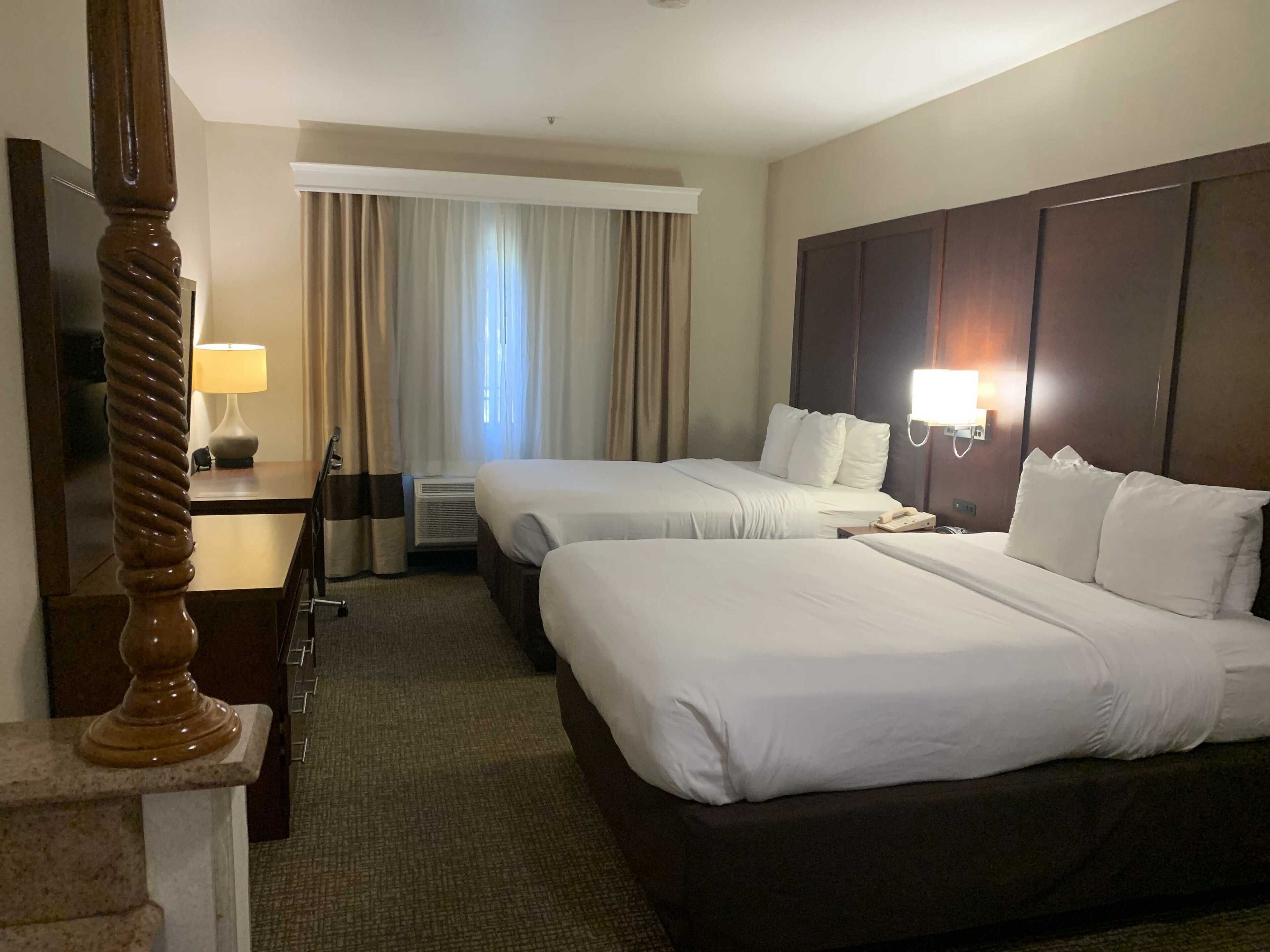 A hotel room with two beds, a desk, a lamp, a telephone, and a window with curtains.